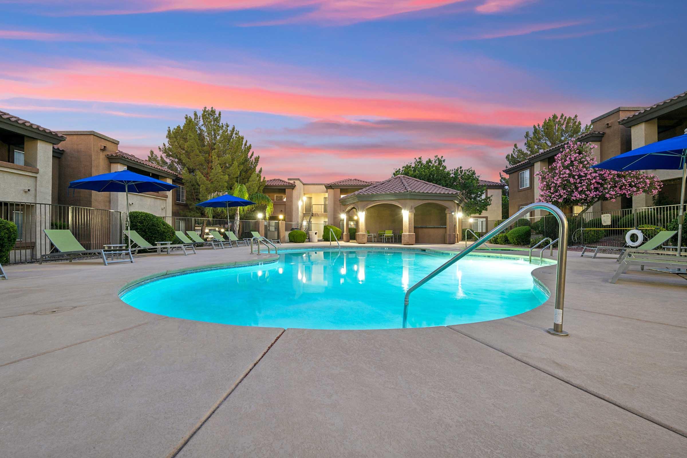 A serene swimming pool surrounded by lounge chairs and blue umbrellas, set against a backdrop of lush greenery and blooming trees.