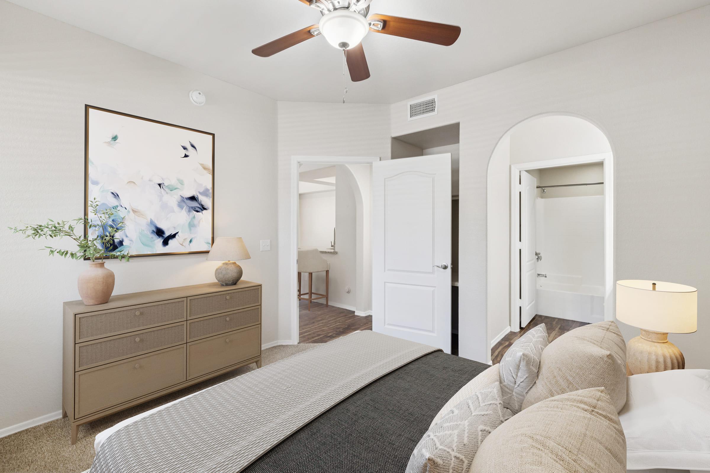 A cozy bedroom featuring a comfortable bed with decorative pillows, a stylish dresser, and a painting of blue flowers on the wall. There's a ceiling fan and doors leading to an adjacent bathroom. The decor is neutral and inviting, creating a warm atmosphere.