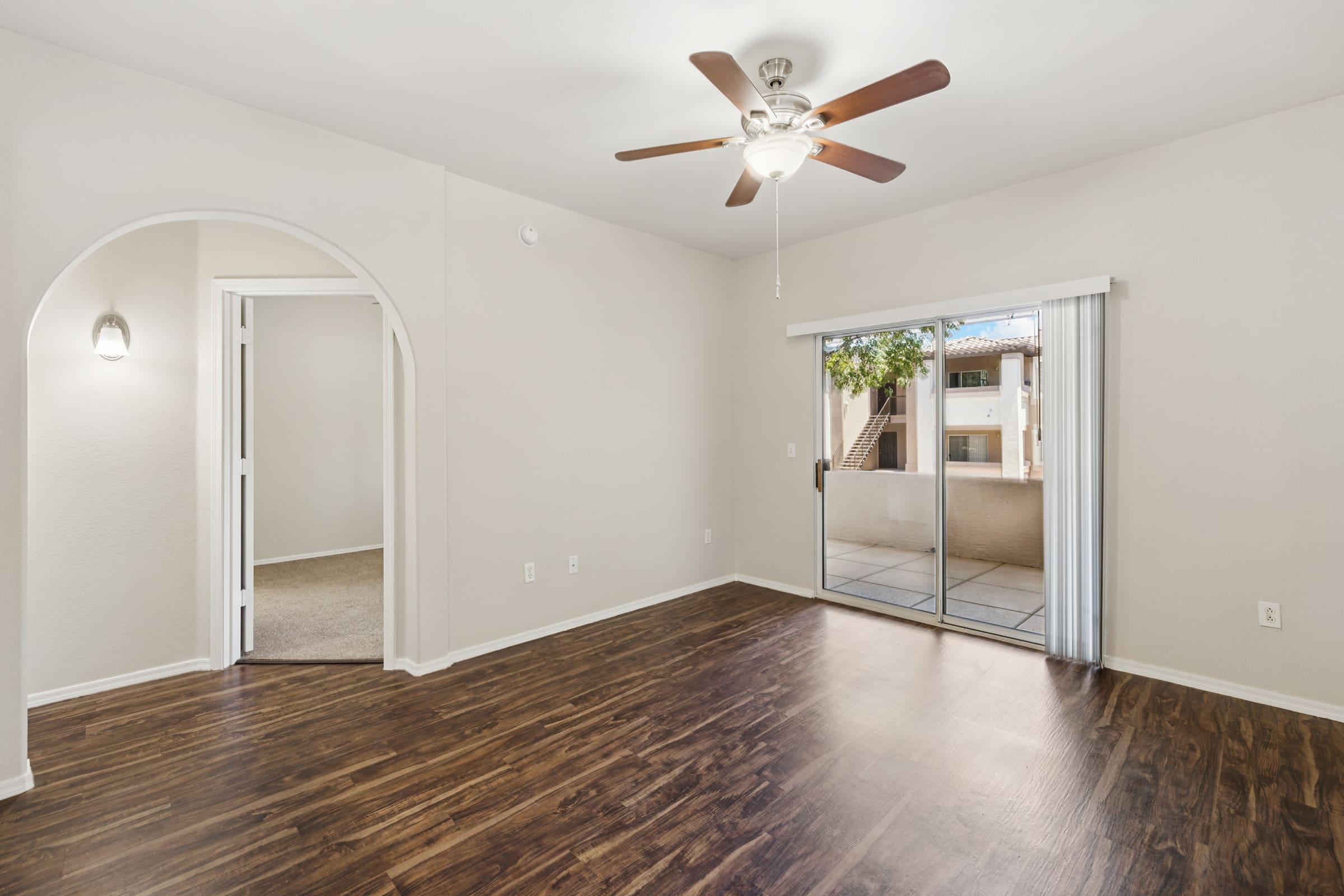 Spacious living room with a ceiling fan, light-colored walls, and dark hardwood flooring. An arched doorway leads to another room, while sliding glass doors provide access to a balcony with outdoor views. Natural light brightens the space, creating an inviting atmosphere.