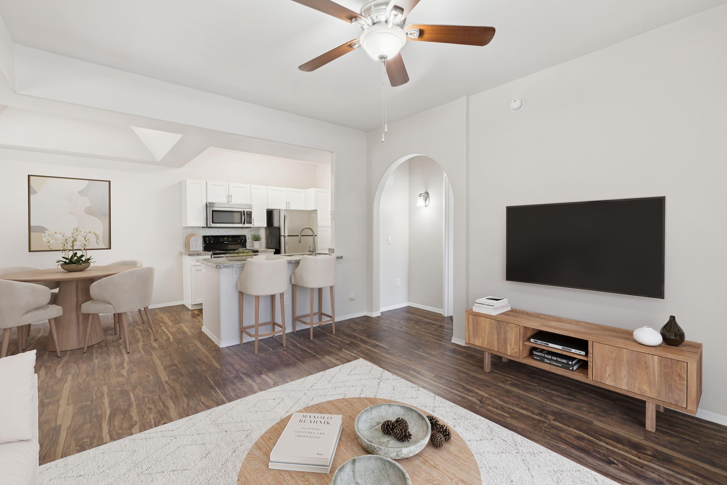 A modern living and dining space featuring a light-colored interior. The living area has a wooden TV stand, a stylish coffee table, and a cozy sofa. The dining area includes a round table with soft chairs. A ceiling fan adds to the airy feel, and the kitchen is visible in the background with white cabinetry and appliances.