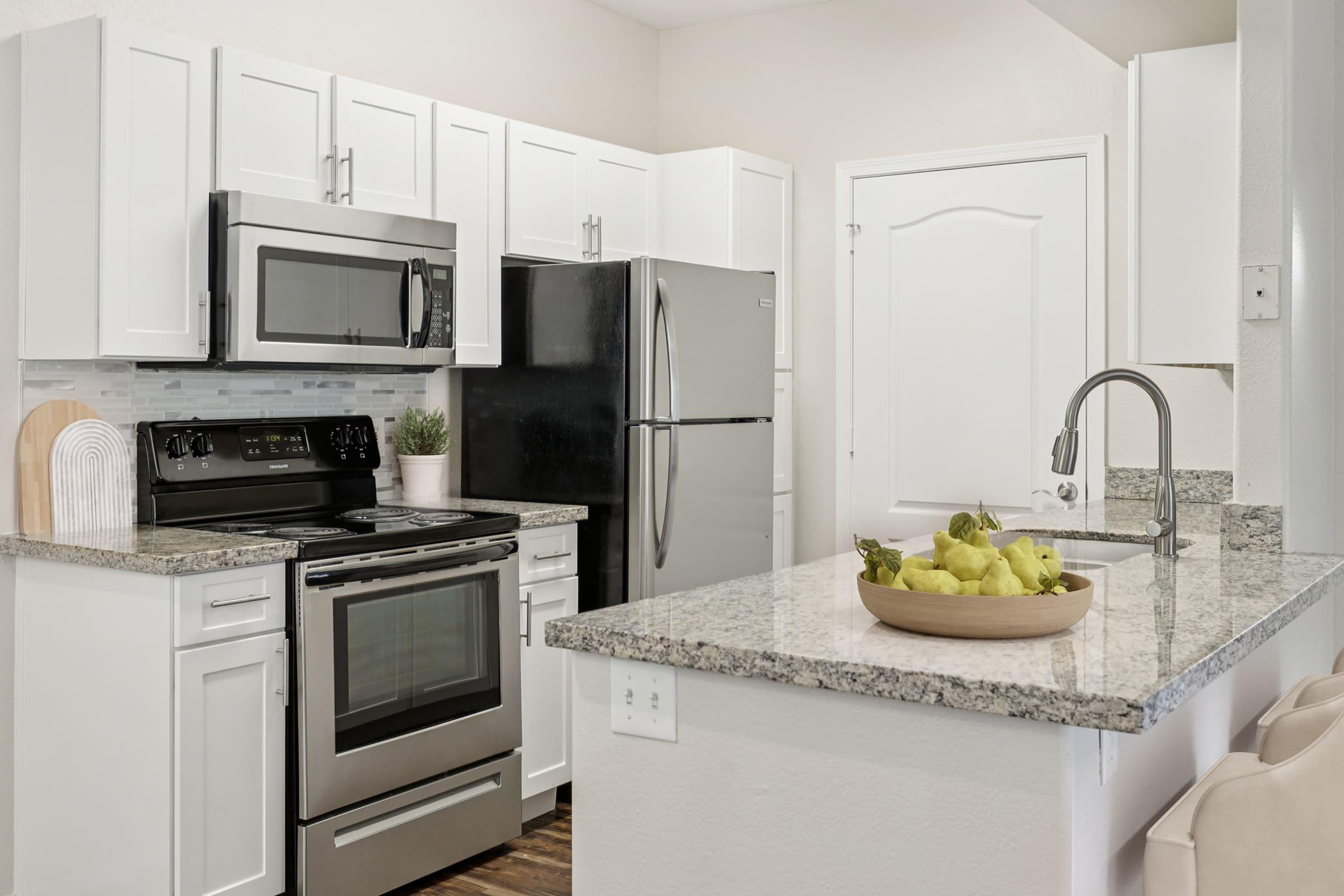 A modern kitchen featuring white cabinets, granite countertops, a stainless steel oven, microwave, and a black refrigerator. A bowl of green pears is placed on the island, and there is a plant on the counter. The room has a bright, inviting atmosphere with neutral tones and ample natural light.