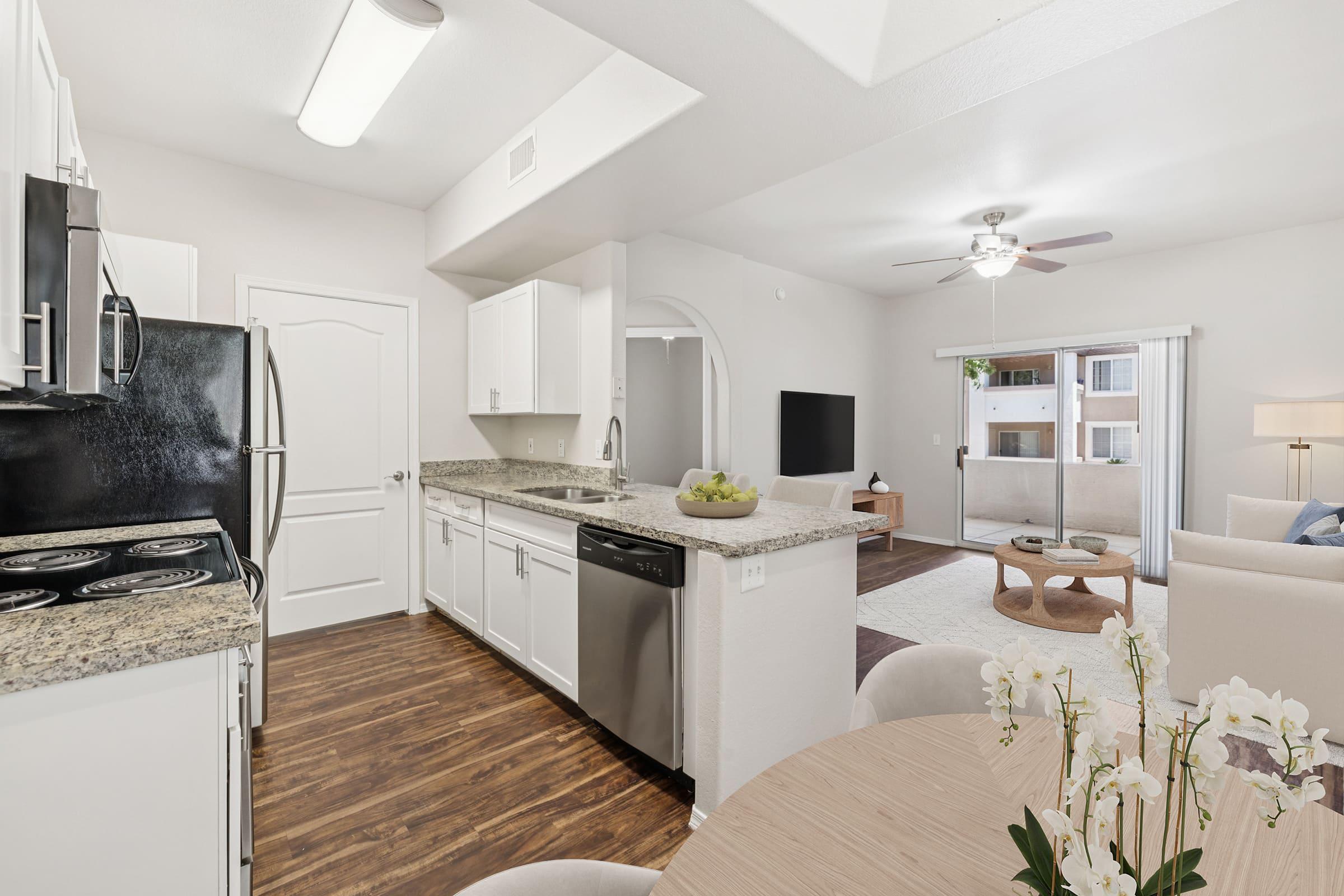 A modern kitchen with white cabinets, granite countertops, and stainless steel appliances. The open layout connects to a living area featuring a ceiling fan and large windows. A dining table with decorative flowers is in the foreground, creating a warm and inviting atmosphere.