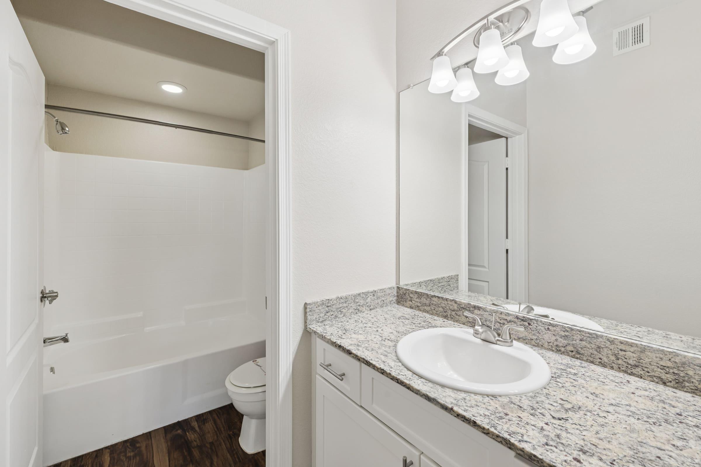 A modern bathroom featuring a granite countertop with a sink, white cabinetry, and a mirror with three lights above. The space includes a bathtub with a shower curtain and a toilet, all set in a bright, neutral-colored room with wood-style flooring.