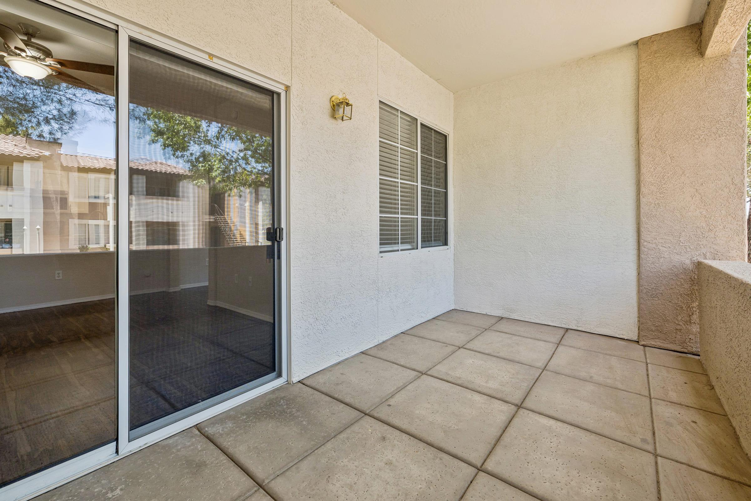 A spacious balcony area with sliding glass doors, surrounded by beige walls. The floor has rectangular tiles, and large windows allow natural light. The view includes part of a building exterior and nearby trees. Ideal for outdoor relaxation or enjoying fresh air.