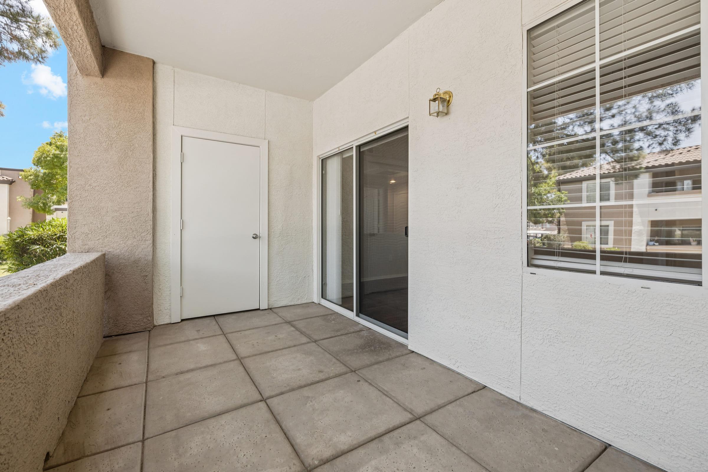 A patio area featuring a smooth tiled floor, a white wall with a door on one side, and a sliding glass door that leads to the interior space. Natural light enters through the sliding door, and there are windows visible on the opposite side, surrounded by greenery.