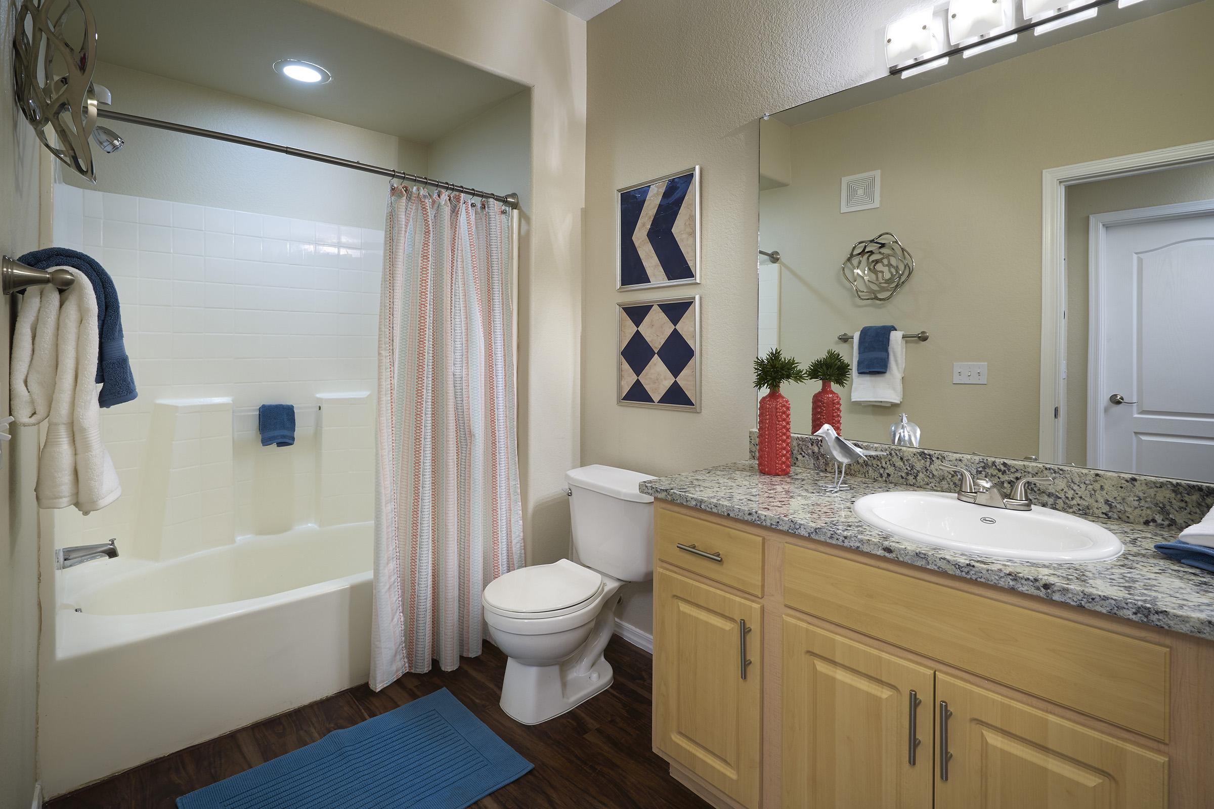 Bright bathroom featuring a shower with a white curtain, a single sink vanity with wooden cabinets, a toilet, and decorative elements like a geometric wall art and small plants.