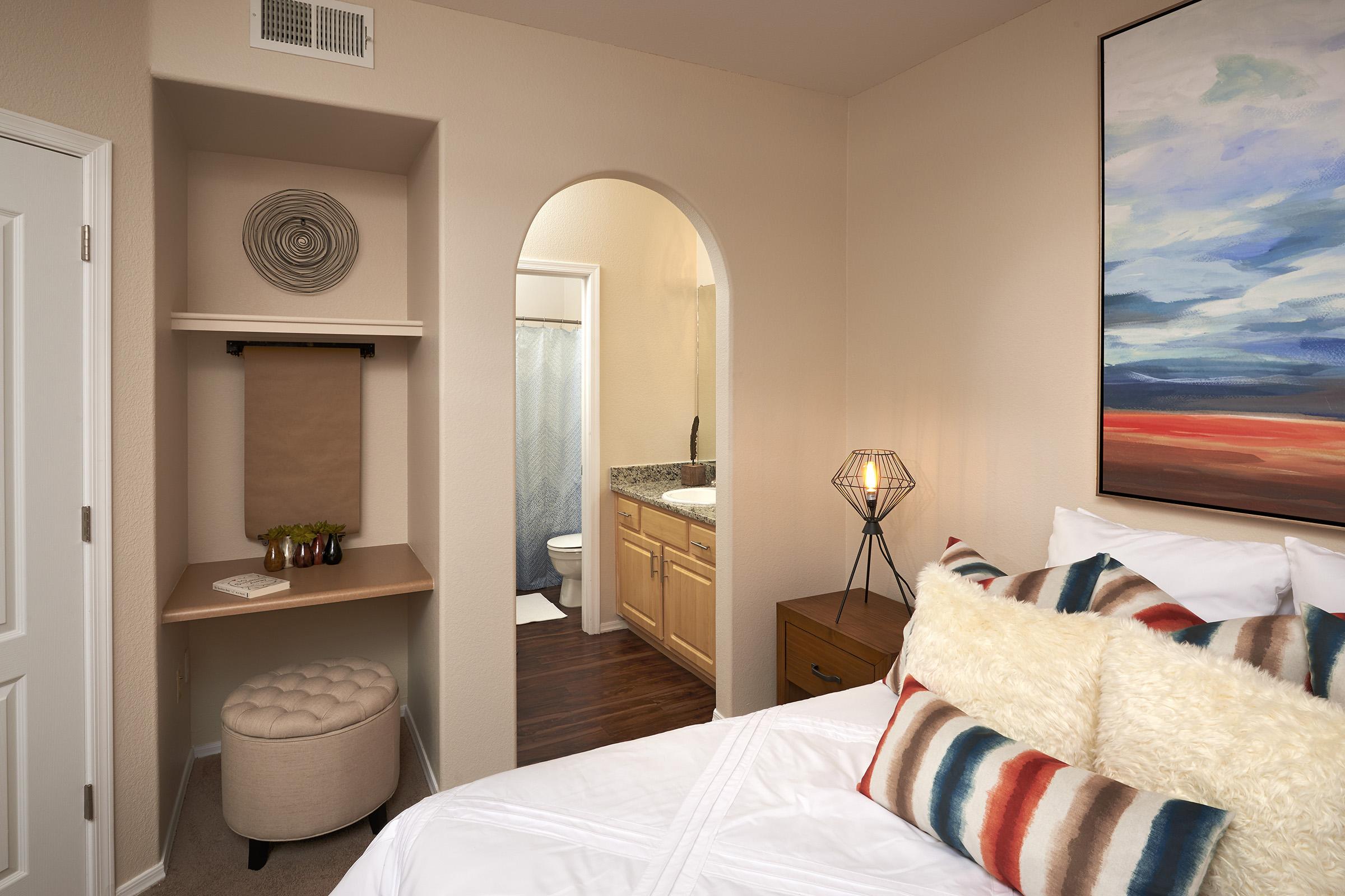 Cozy bedroom featuring a large bed with decorative pillows, a stylish lamp, and a small seating area. An alcove with a shelf and an archway leads to a bathroom with a shower.