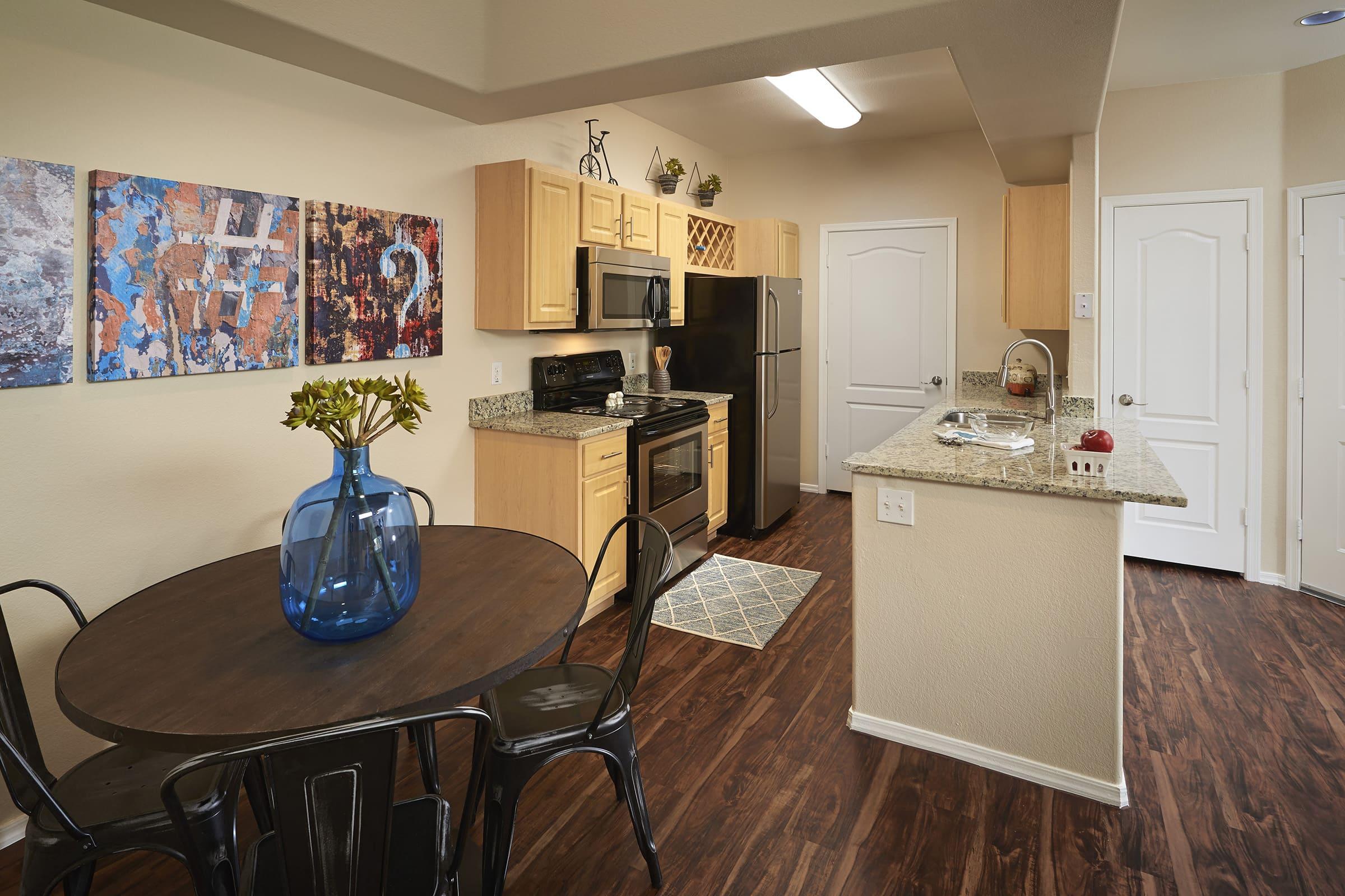 Contemporary kitchen with wooden cabinets and granite countertops, featuring a dining table with a blue vase and black chairs. Appliances include a microwave, oven, and fridge.