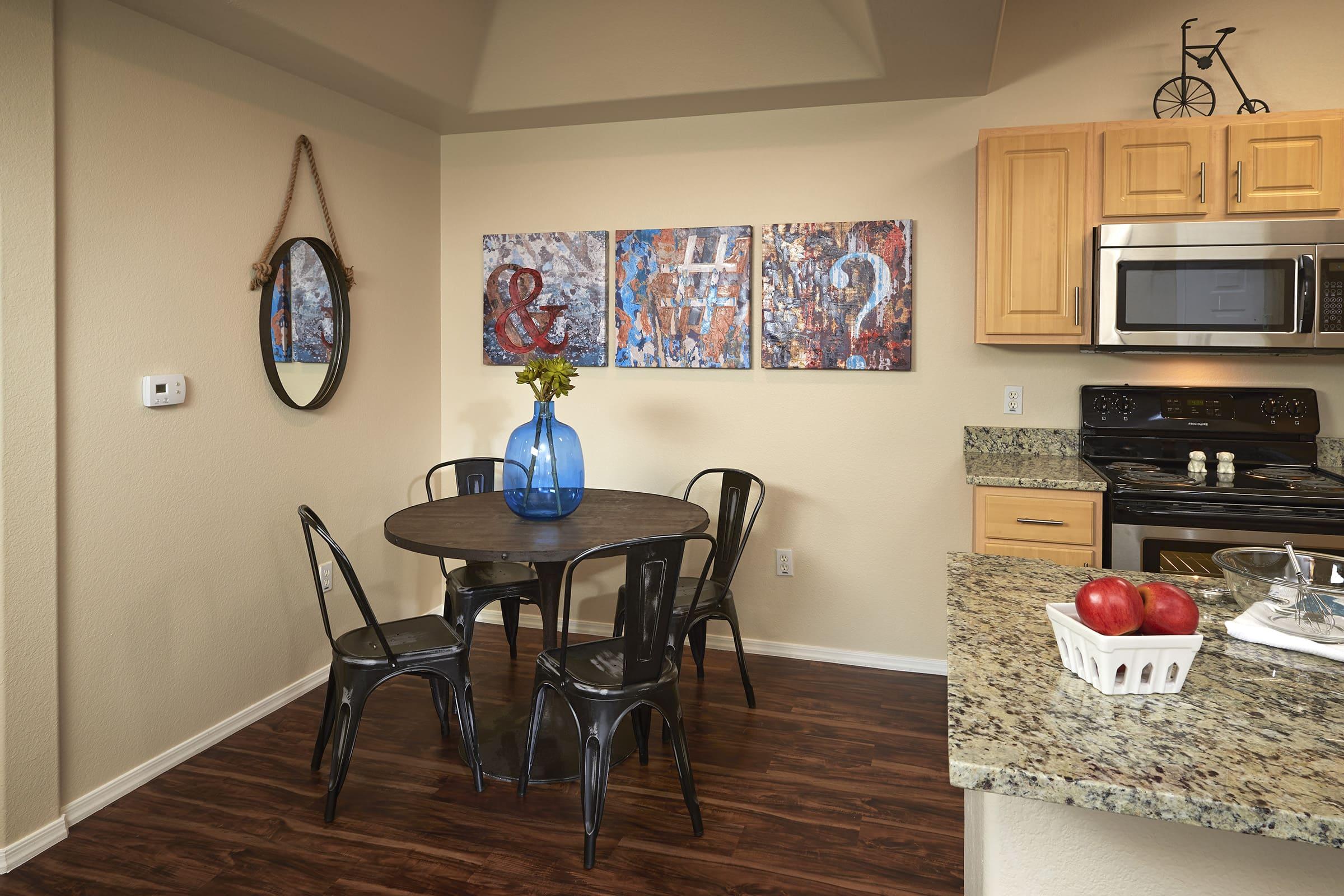 A modern kitchen with light wooden cabinets, a granite countertop, and a small dining area featuring a round black table surrounded by metal chairs.