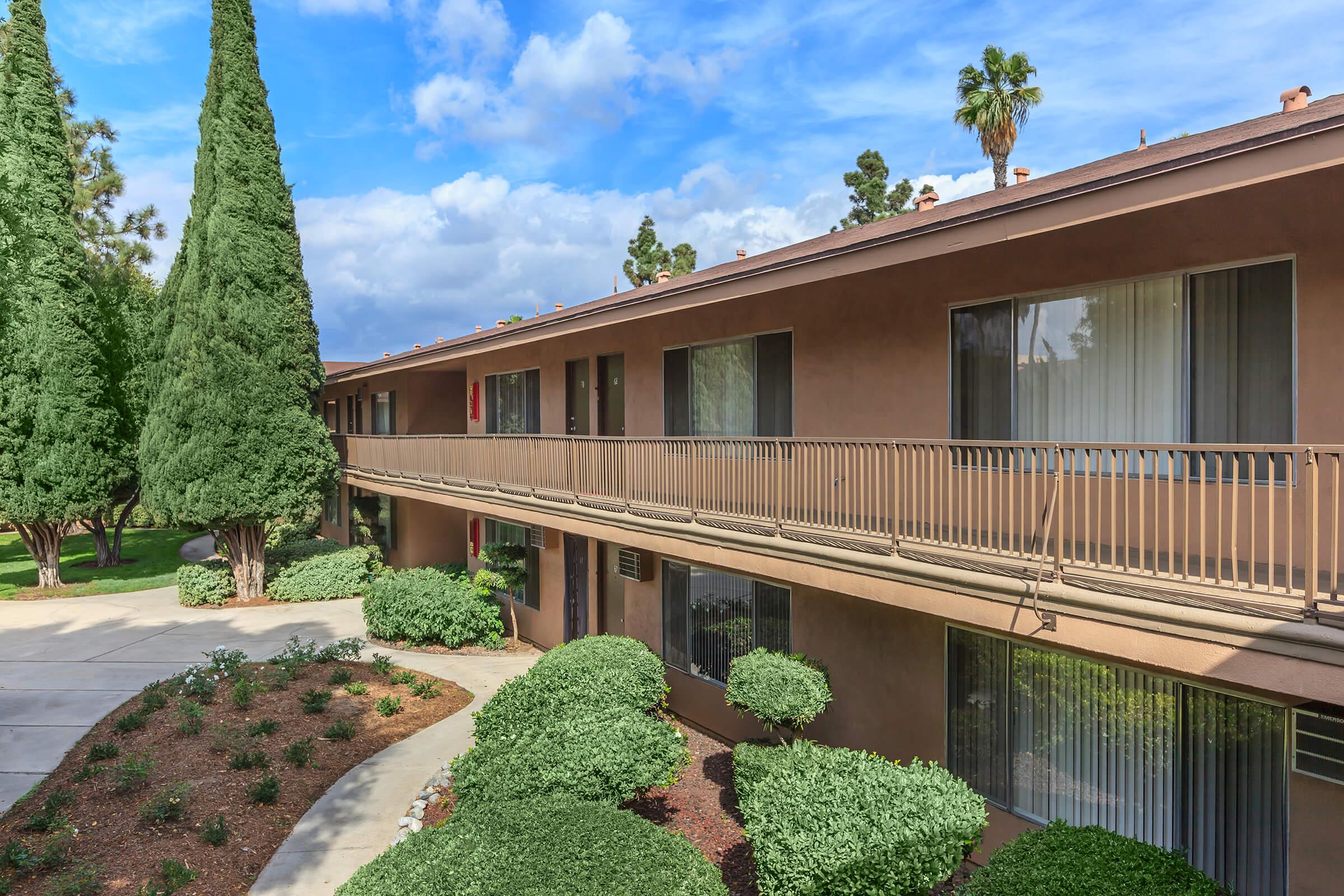A view of a two-story building with a balcony overlooking a landscaped area. The exterior features large windows and is surrounded by neatly trimmed shrubs and palm trees. The sky is partly cloudy, creating a serene atmosphere.