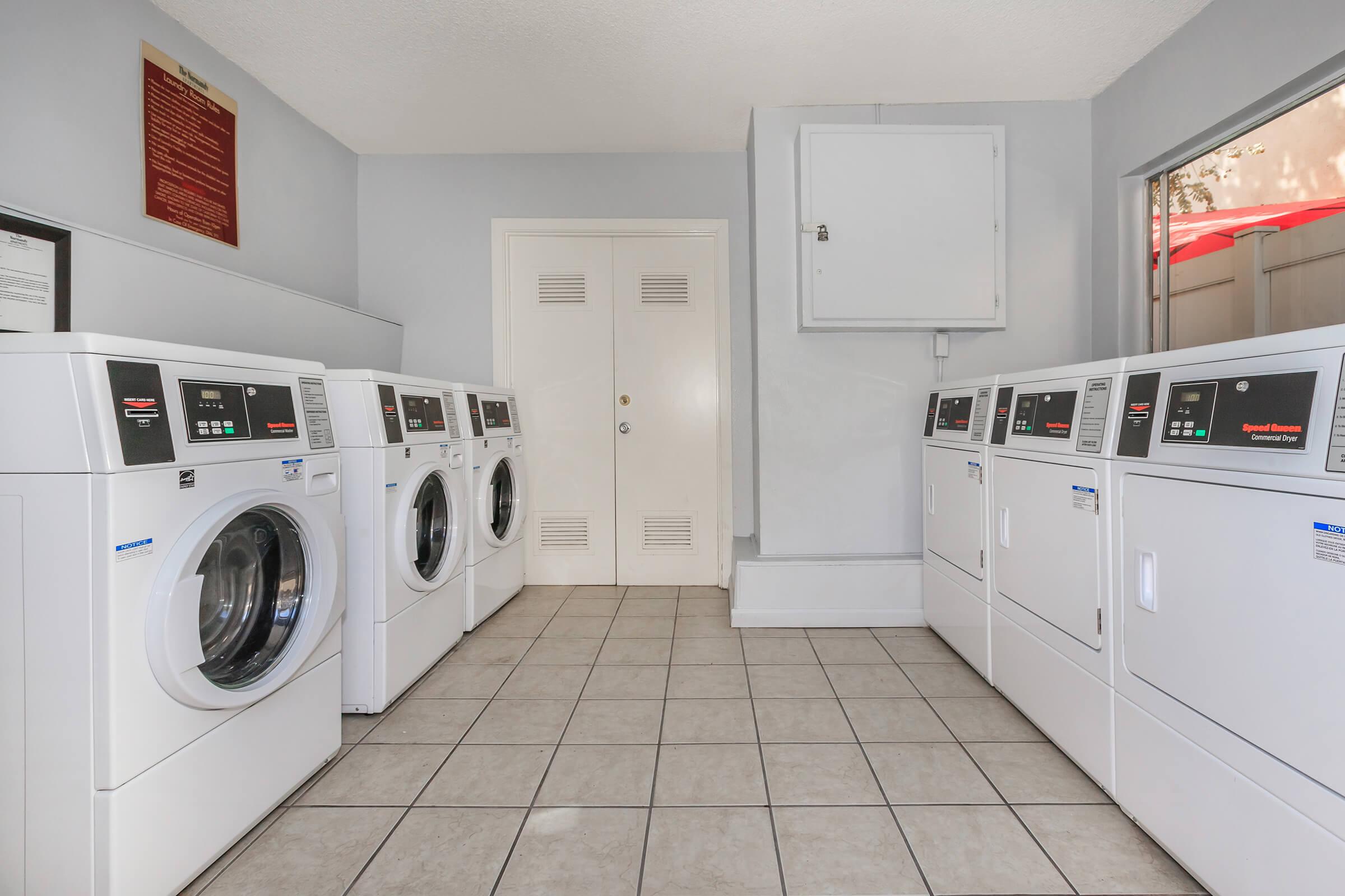 A clean and modern laundry room featuring several white washing machines and dryers arranged in a row. The floor is tiled, and the walls are painted light gray. There is a door on one side and an information board mounted on the wall. Natural light is coming in from a window, creating a bright atmosphere.