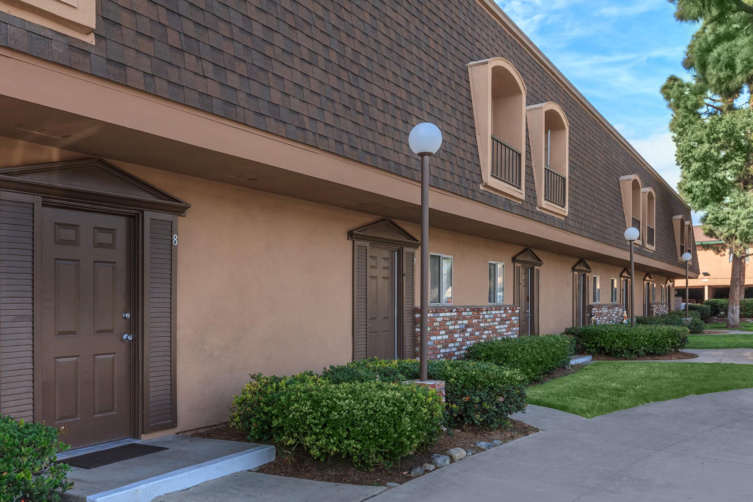A row of residential units with doors and windows, featuring a brown shingle-style exterior. Each unit has a small porch area, with landscaped bushes and grass in the foreground. Street lamps are positioned along the walkway, creating a well-maintained appearance in a sunny setting.
