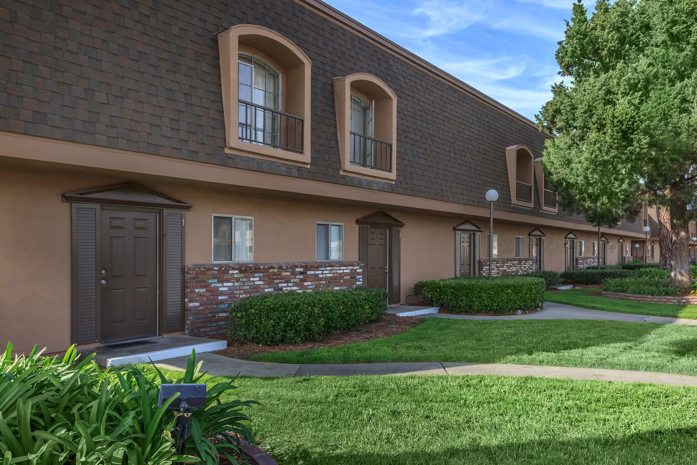 A well-maintained residential area featuring a row of townhouses with brown shingle siding and brick accents. Each unit has a front door with a small overhang and balconies above. Lush green landscaping surrounds the buildings, including shrubs and well-manicured grass, under a clear blue sky.