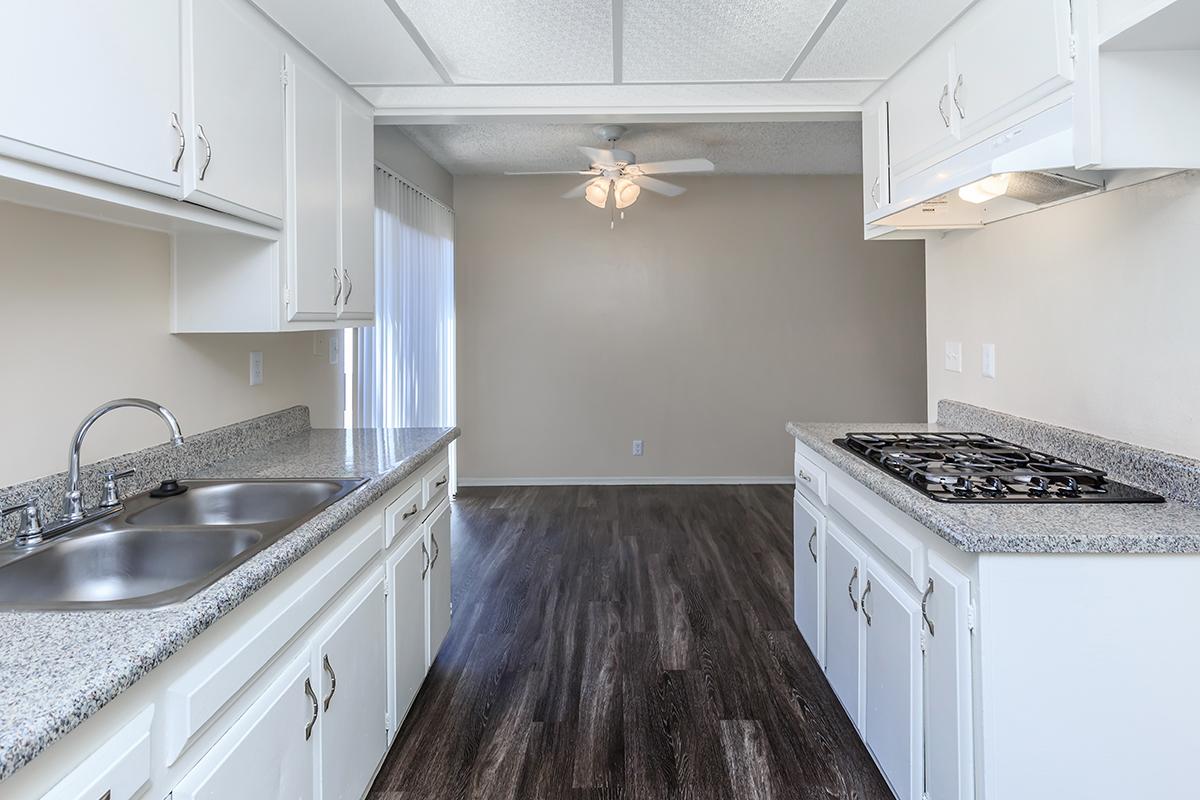 A modern kitchen featuring white cabinets, a double sink, and a gas stove with a granite countertop. The space has a ceiling fan and light-colored walls, with an open layout leading into a living area with wood-style flooring. Large windows provide natural light.