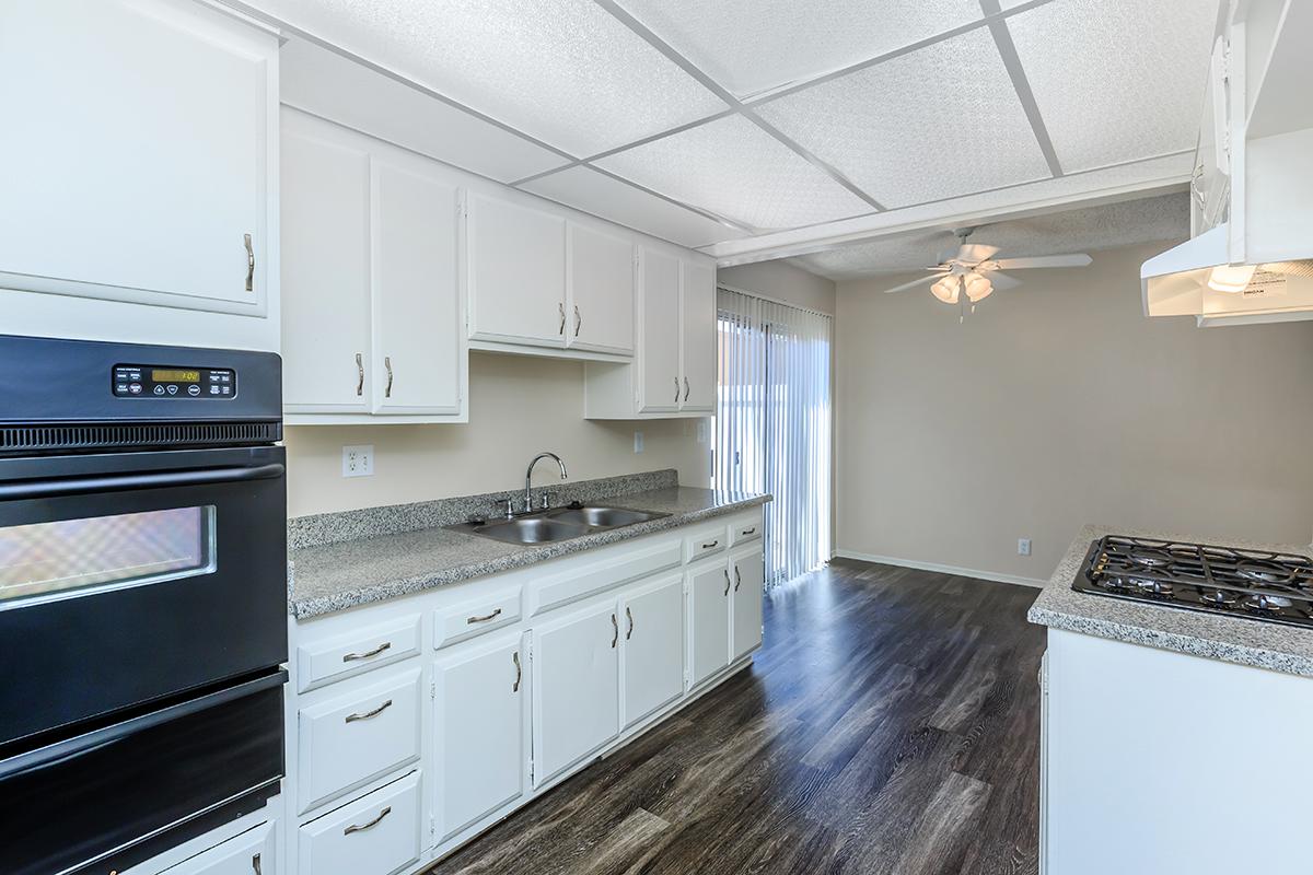 A bright kitchen featuring white cabinetry, a granite countertop, and a black oven. There's a gas stove on the right, and a window with blinds allows natural light. The floor has a dark, modern finish, and the adjacent dining area is visible, complete with a ceiling fan and beige walls.