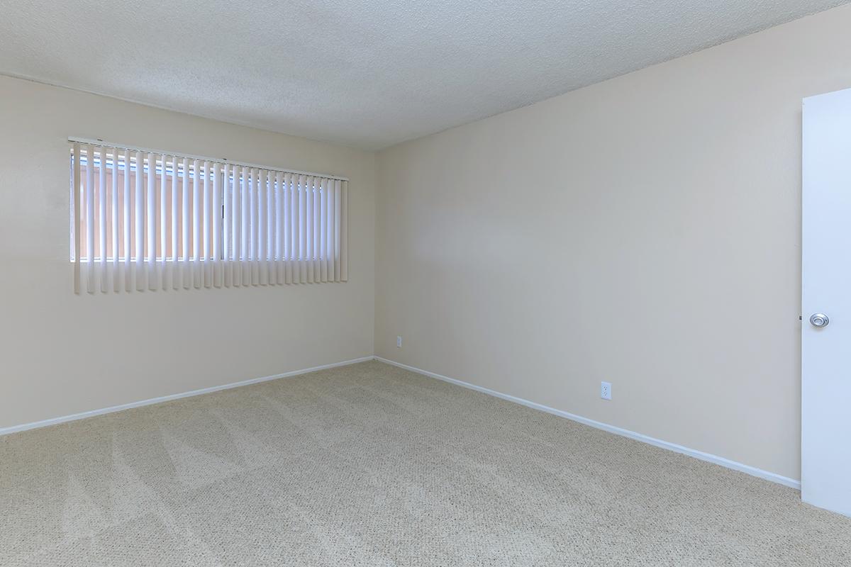 Empty room with light beige walls and carpet. A window with horizontal blinds allows natural light to enter. The space has minimal furnishings, featuring a single door on the right side. Overall, the room appears clean and well-maintained, ready for occupancy or decoration.