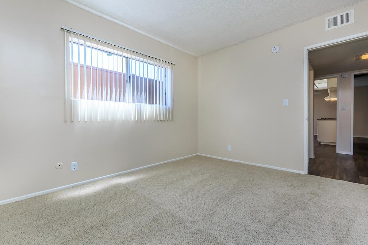 Empty room with beige walls and carpet, featuring a window with vertical blinds letting in natural light. A doorway on the right leads to another space. The room has minimal furnishings, creating a spacious and clean appearance.
