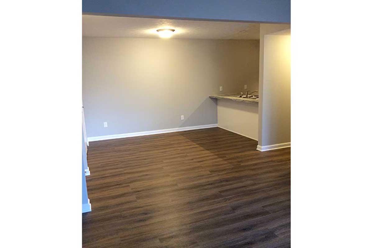A spacious, empty room featuring light brown wooden flooring. The walls are painted in a soft gray, and there is a ceiling light fixture. To the right, a kitchen area is partially visible with a countertop and open space. The overall atmosphere is clean and ready for furnishing.