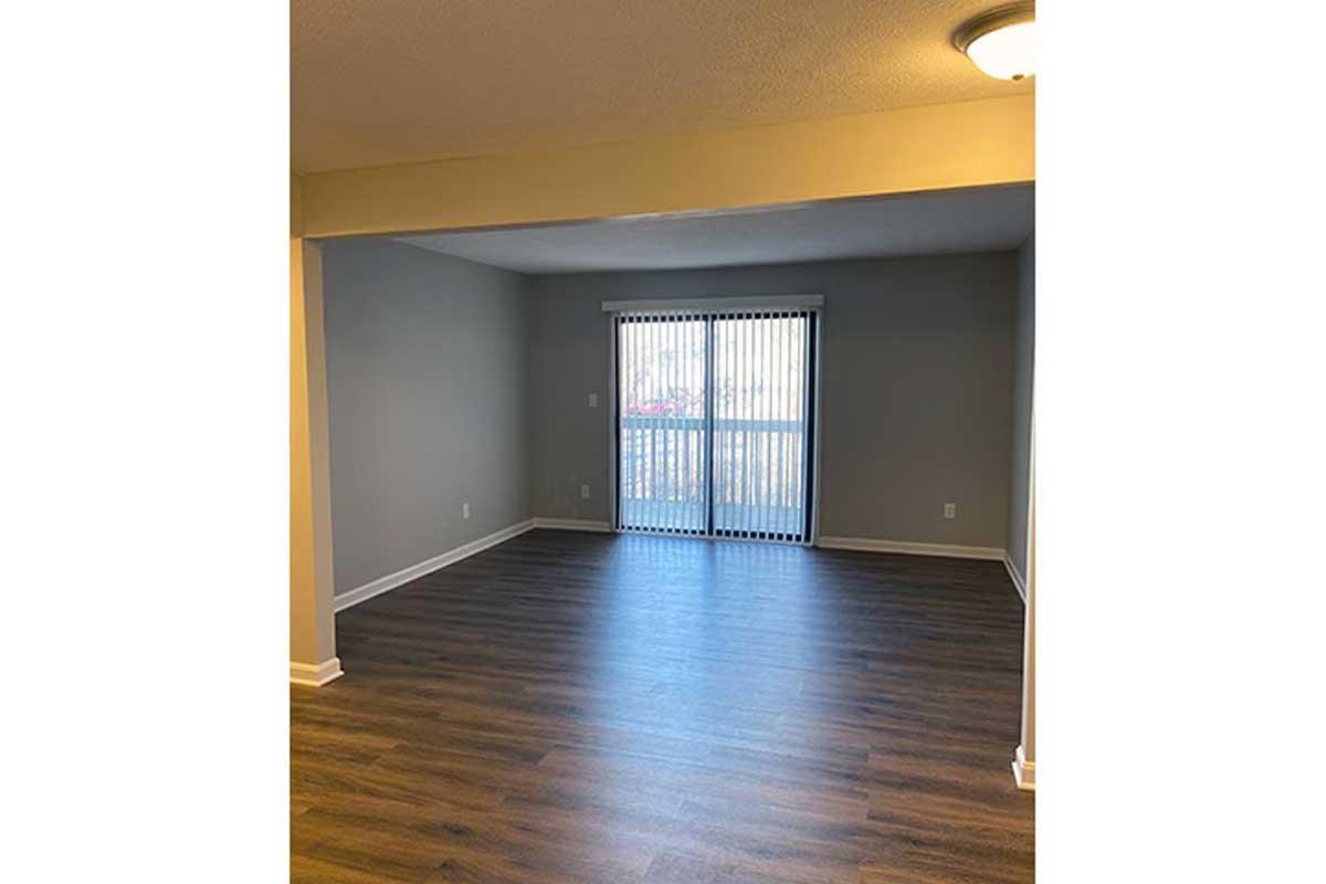 A spacious, empty living room with light gray walls and a wood laminate floor. A sliding glass door with vertical blinds leads to a balcony, allowing natural light to enter the room. The ceiling features a light fixture, and there are no furniture or decorations present.