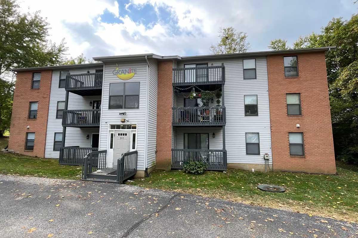 A three-story apartment building featuring a mix of brick and siding, with balconies on each floor. The building has a central entrance, and the name "Granite" is displayed above the door. Surrounding trees and a paved pathway are visible, along with fallen leaves on the ground.