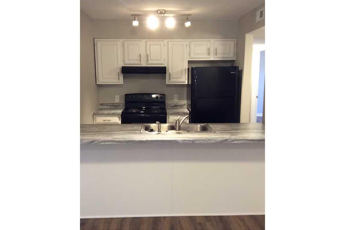 A modern kitchen with white cabinets and countertops, featuring an electric stove, a black refrigerator, and a sink with a dual faucet. The space is well-lit with recessed lighting above and has an open layout leading to another room. The flooring appears to be a wood laminate.