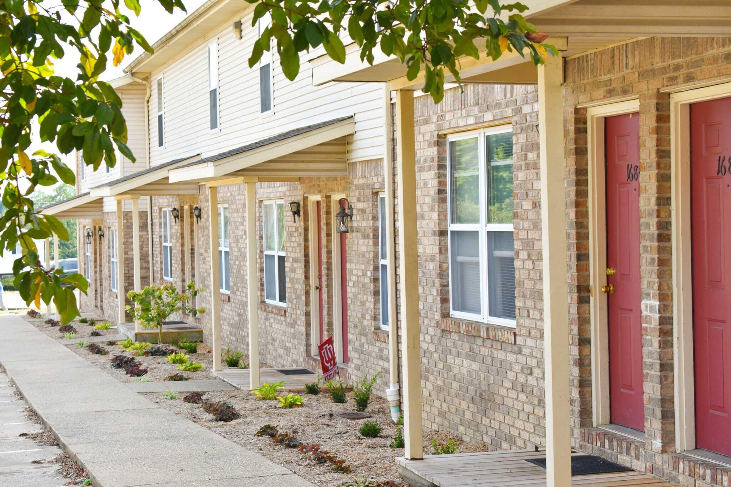 Row of brick townhouses with red doors, each featuring a covered porch. The walkway is lined with small plants and shrubs, and a few trees are partially visible, providing a touch of greenery against the exterior of the buildings.