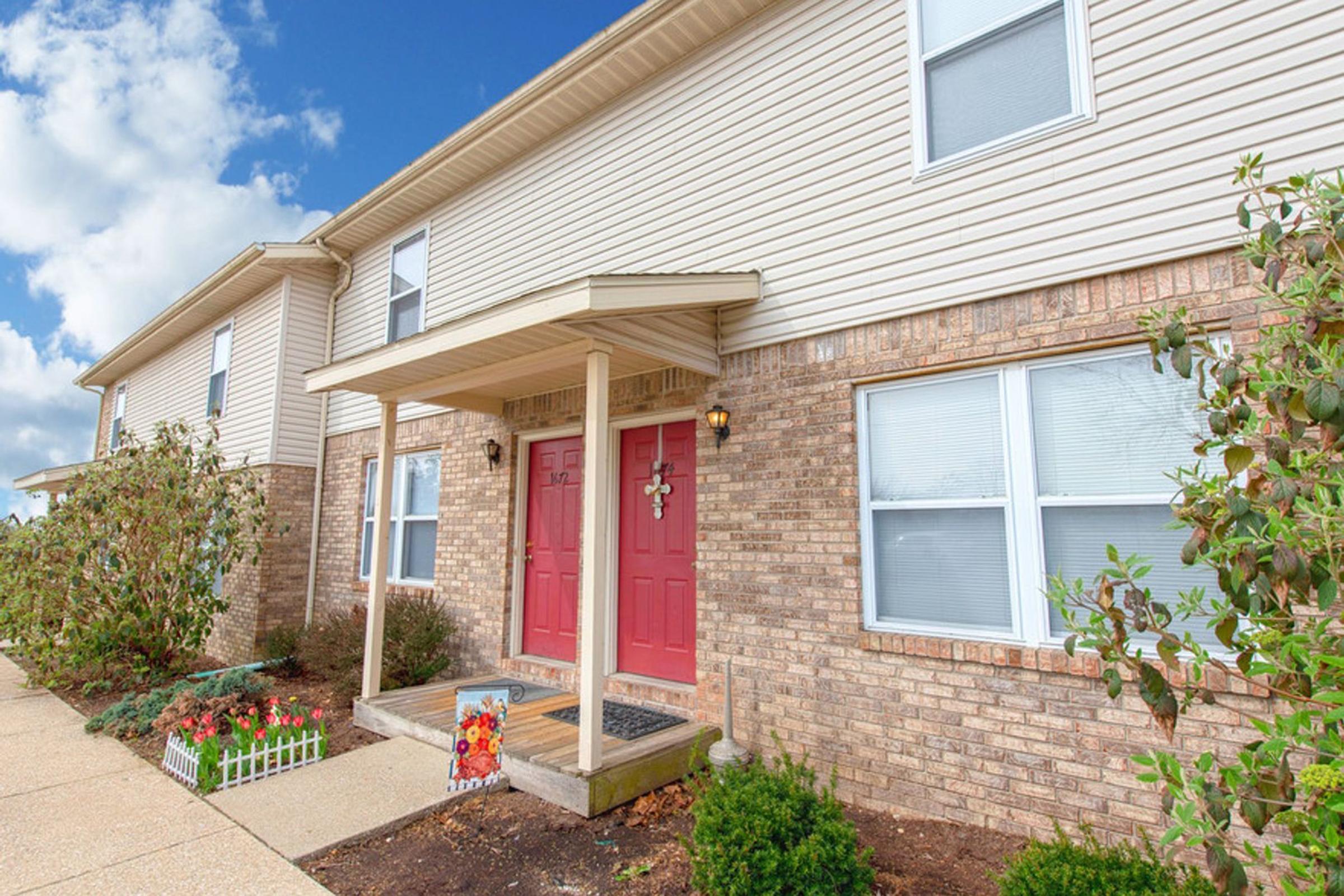 Exterior view of a two-story residential building featuring beige siding and a brick facade. The entrance has a covered porch with double red doors. Flanking the entrance are small bushes and a decorative sign on the ground. The sky is partly cloudy, adding a bright ambiance to the scene.