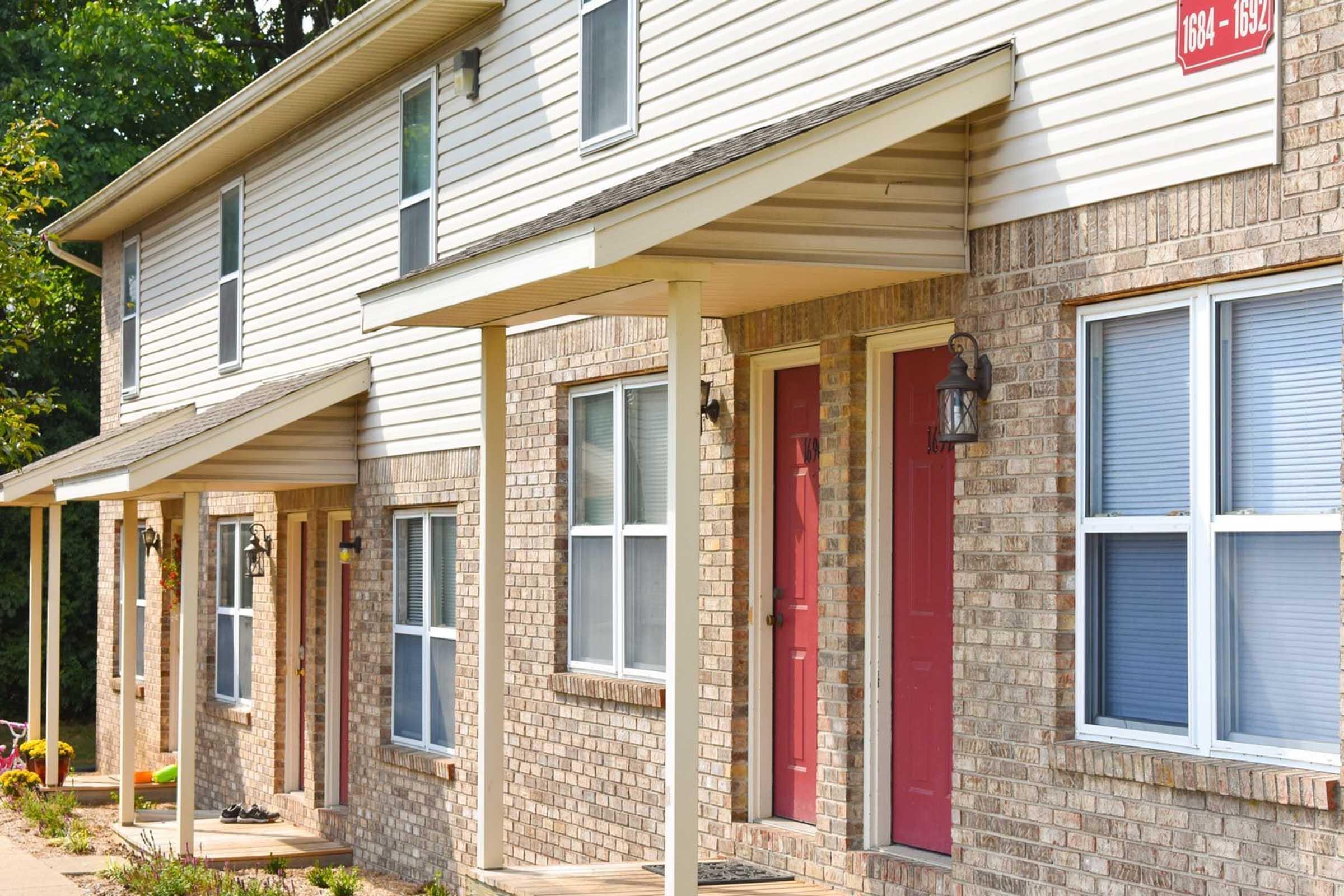 A row of townhouses featuring light-colored siding and brick facades. Each unit has a red door and covered entryway, with windows on either side. The scene is set in a well-maintained environment with greenery in the background.