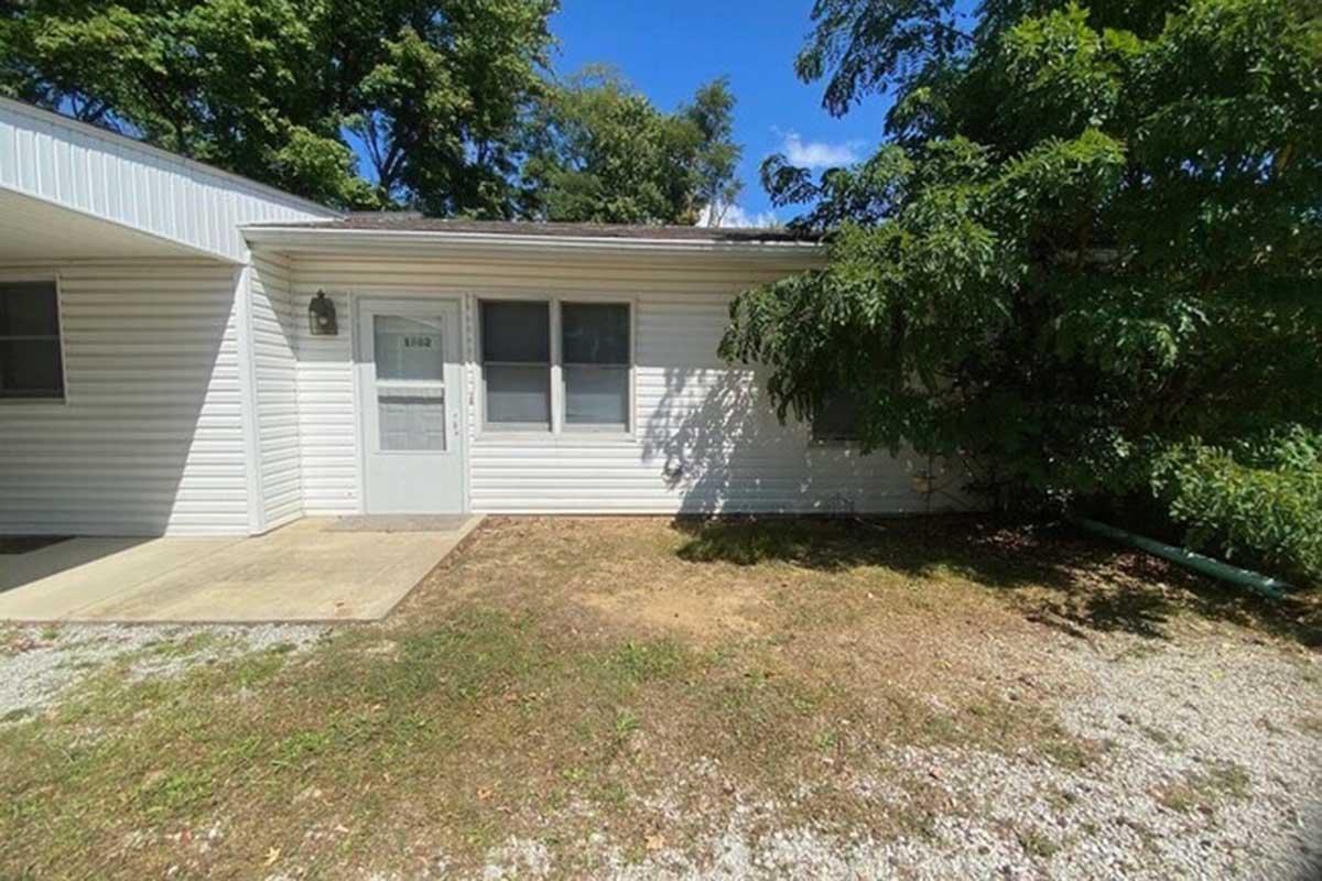 Exterior of a single-story house featuring a white siding facade, a front door with a window, and a small porch area. The surrounding yard has grass and a few trees providing shade, with a clear blue sky overhead.
