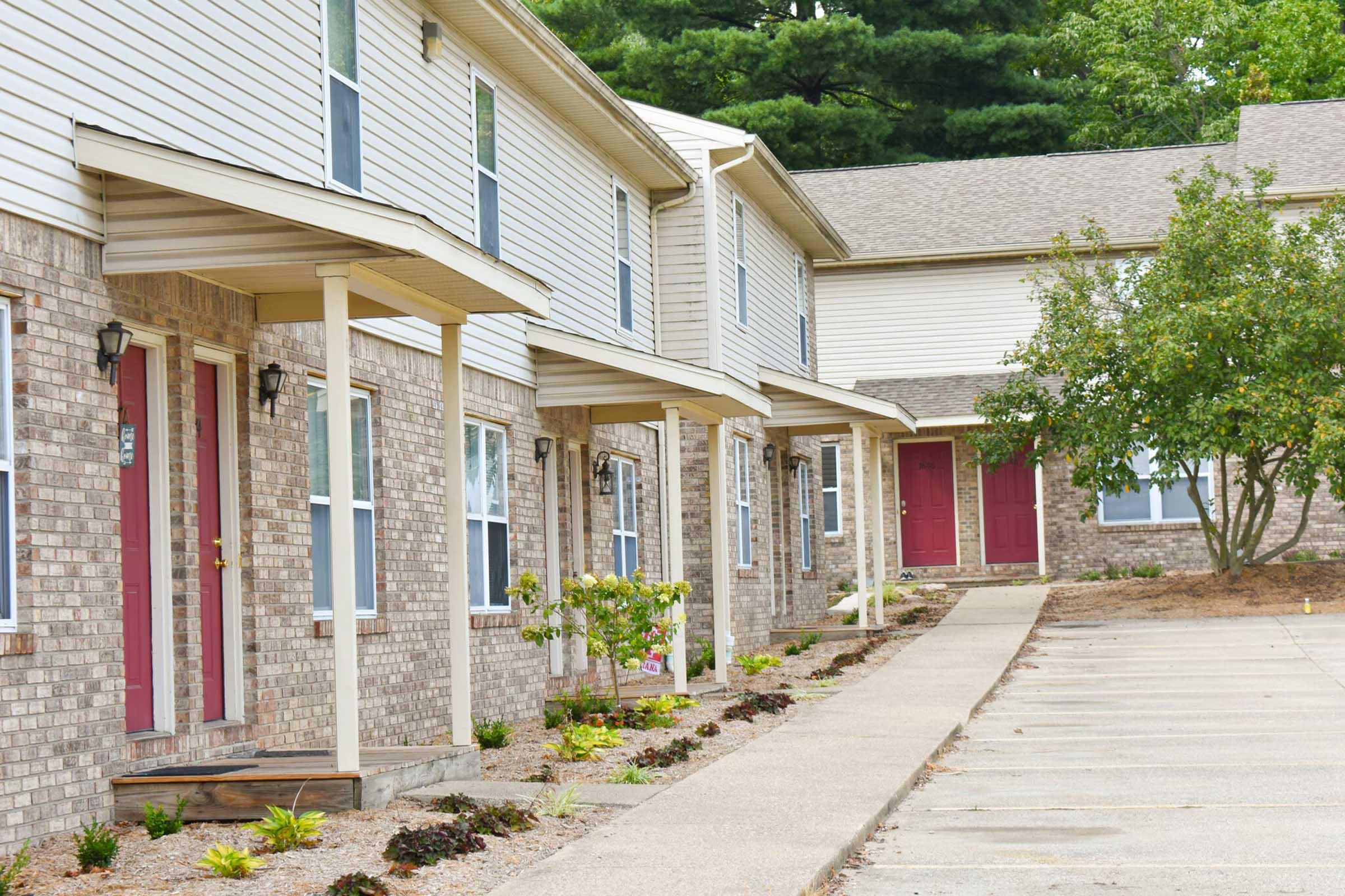 A row of brick townhouses with four units, each featuring a front porch and decorative landscaping. The doors are different shades of red and there are shrubs and plants along the walkway. The scene is set in a quiet residential area surrounded by trees.