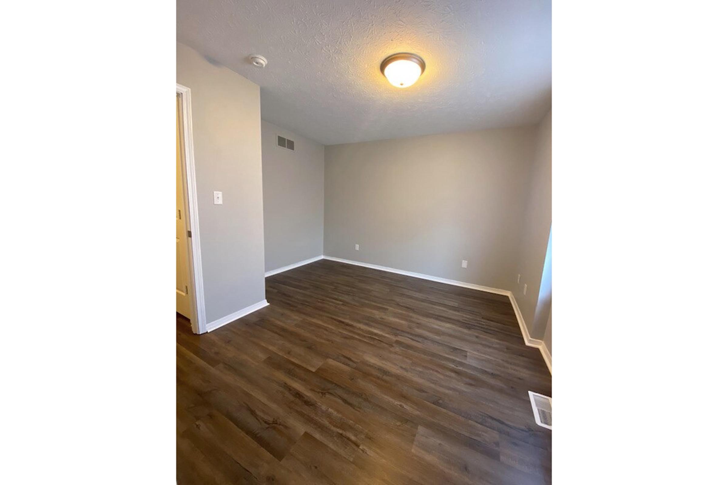 Empty room with light gray walls and a wooden floor. The ceiling features a round light fixture. There is a single door visible on the left side leading to another room. The space is well-lit, creating an open and inviting atmosphere. No furniture or decorations are present.