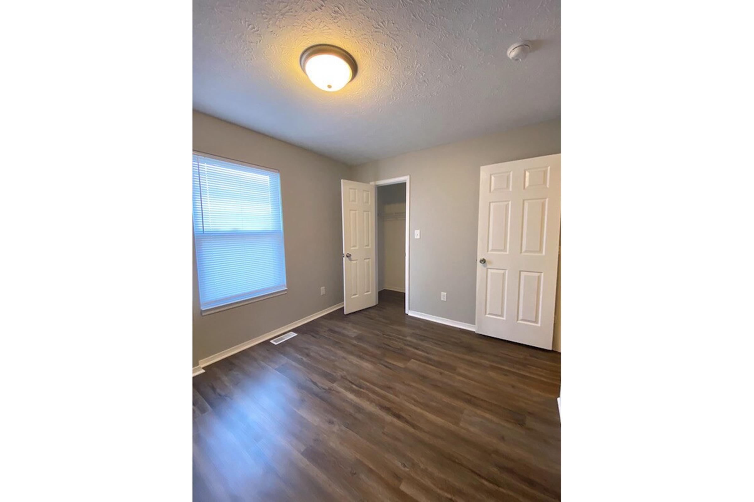 A neutral-colored room with light gray walls and wooden flooring. There is a ceiling light fixture, a window with light blinds, and two closed doors on the right side. The room is empty, creating a spacious feel.