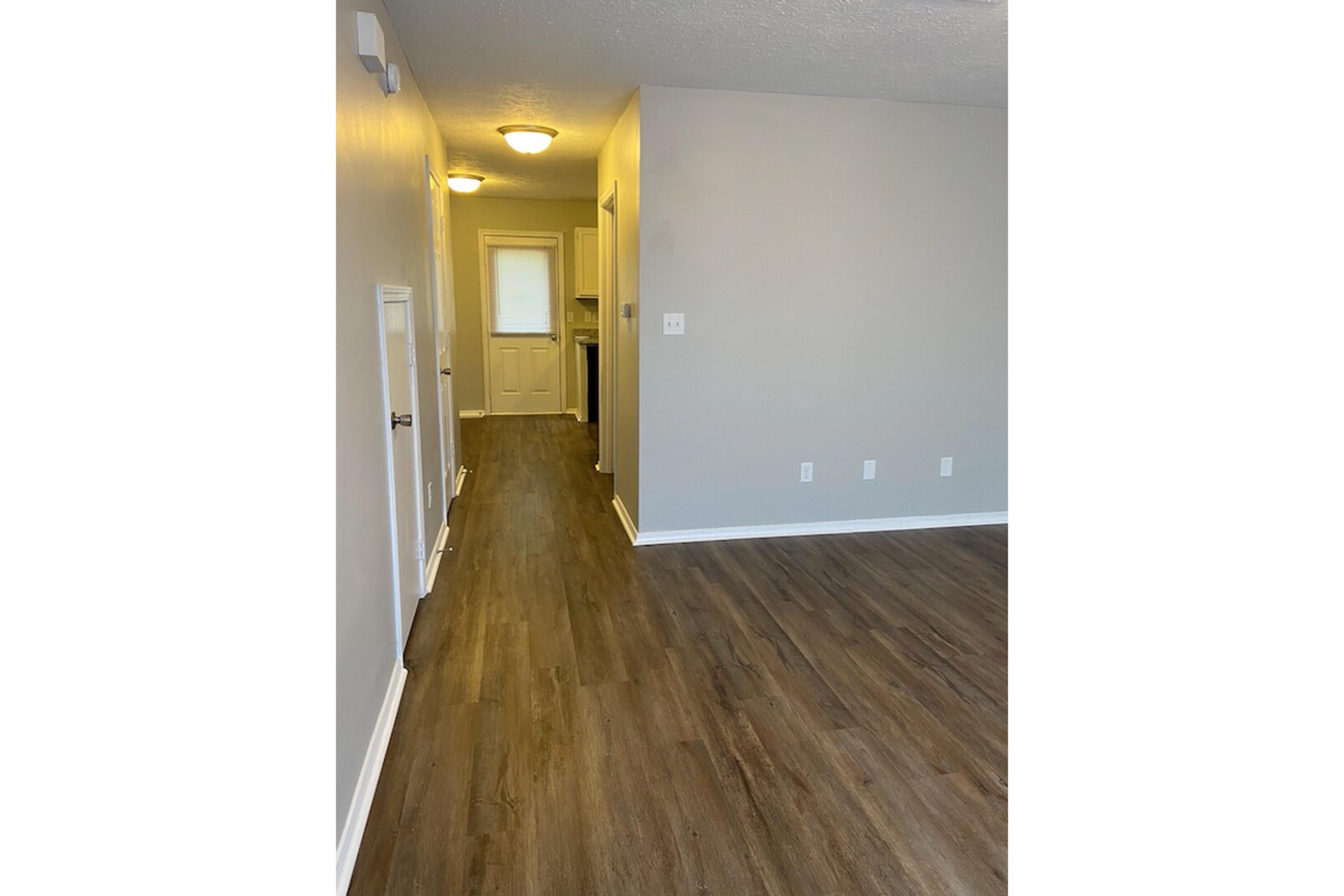 A hallway in a residential space featuring light gray walls, new flooring, and a white door at the end. The area is well-lit with ceiling lights and includes a small closet door on the left. The overall aesthetic is modern and clean, suggesting a fresh and inviting atmosphere.
