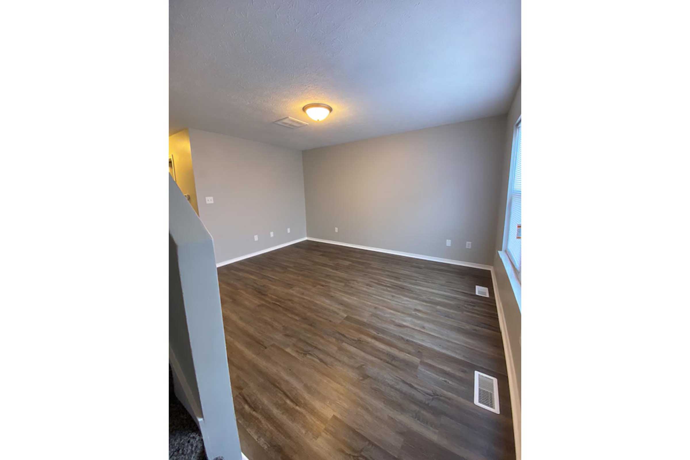 Empty room with light gray walls and new wooden flooring. It features a ceiling light fixture and a window allowing natural light. The space is uncluttered, with minimal decor and no furniture, creating a bright and open atmosphere.