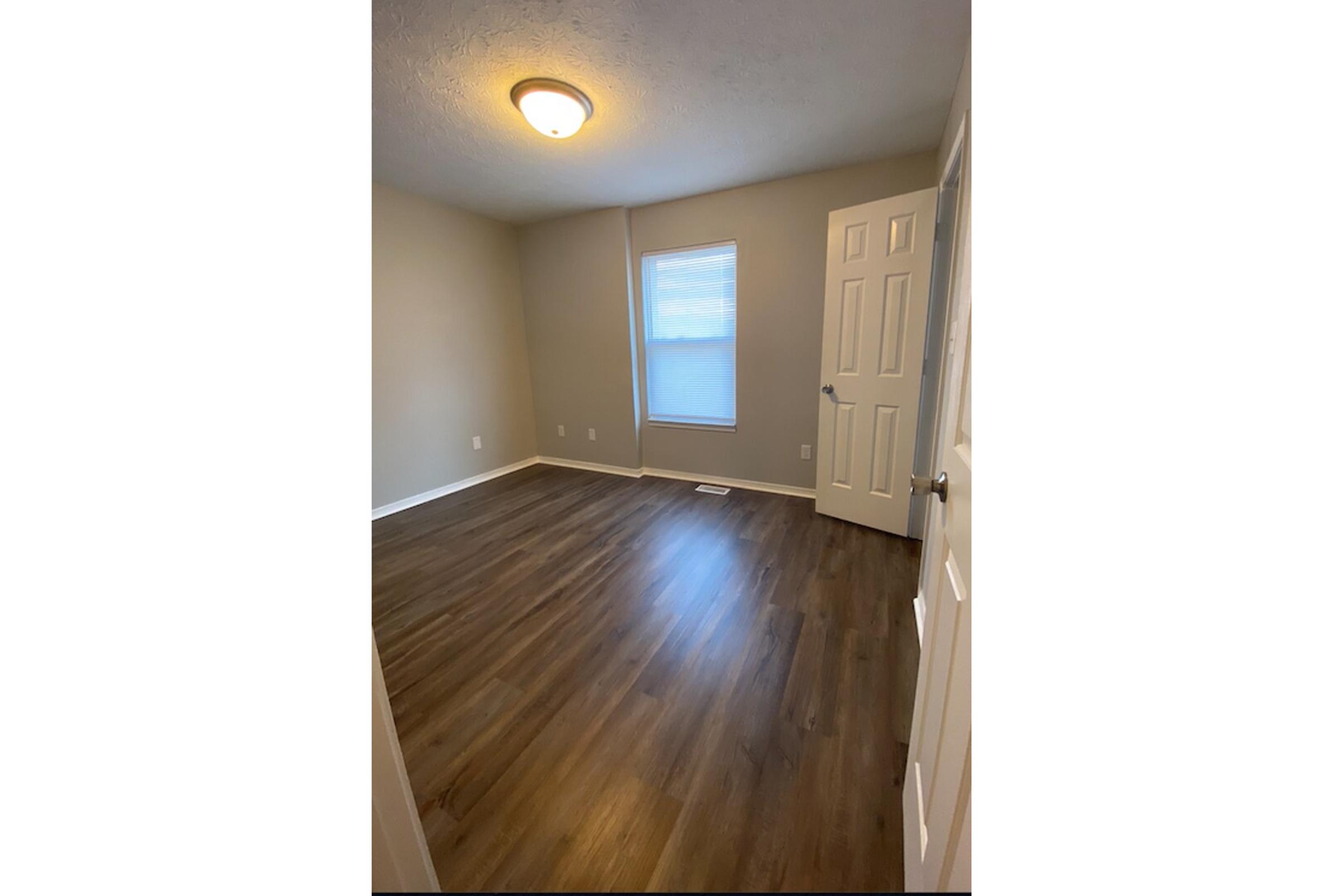 A spacious, empty room featuring light-colored walls, a ceiling light fixture, and a window with blinds. The floor is made of hardwood or laminate, and there is a door on the right leading to another area. The overall atmosphere is bright and neutral, suitable for various uses.