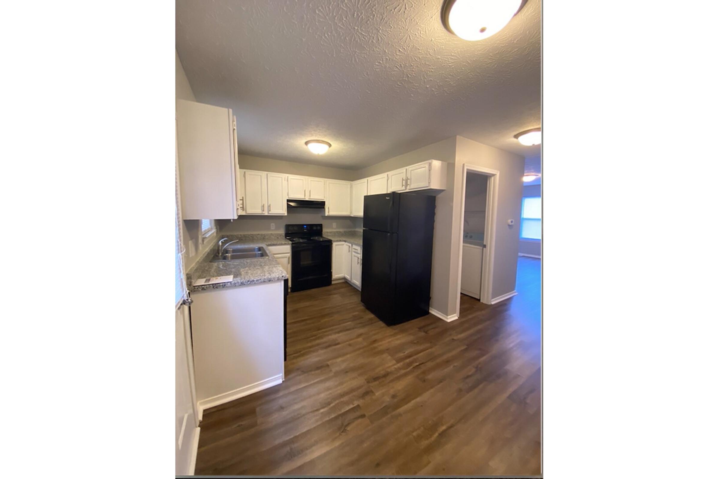 A modern kitchen featuring white cabinetry, granite countertops, and stainless steel appliances. The space includes a black refrigerator and stove, with a view of an adjoining room. The floor is hardwood, and there's overhead lighting for illumination. Natural light enters through a window.