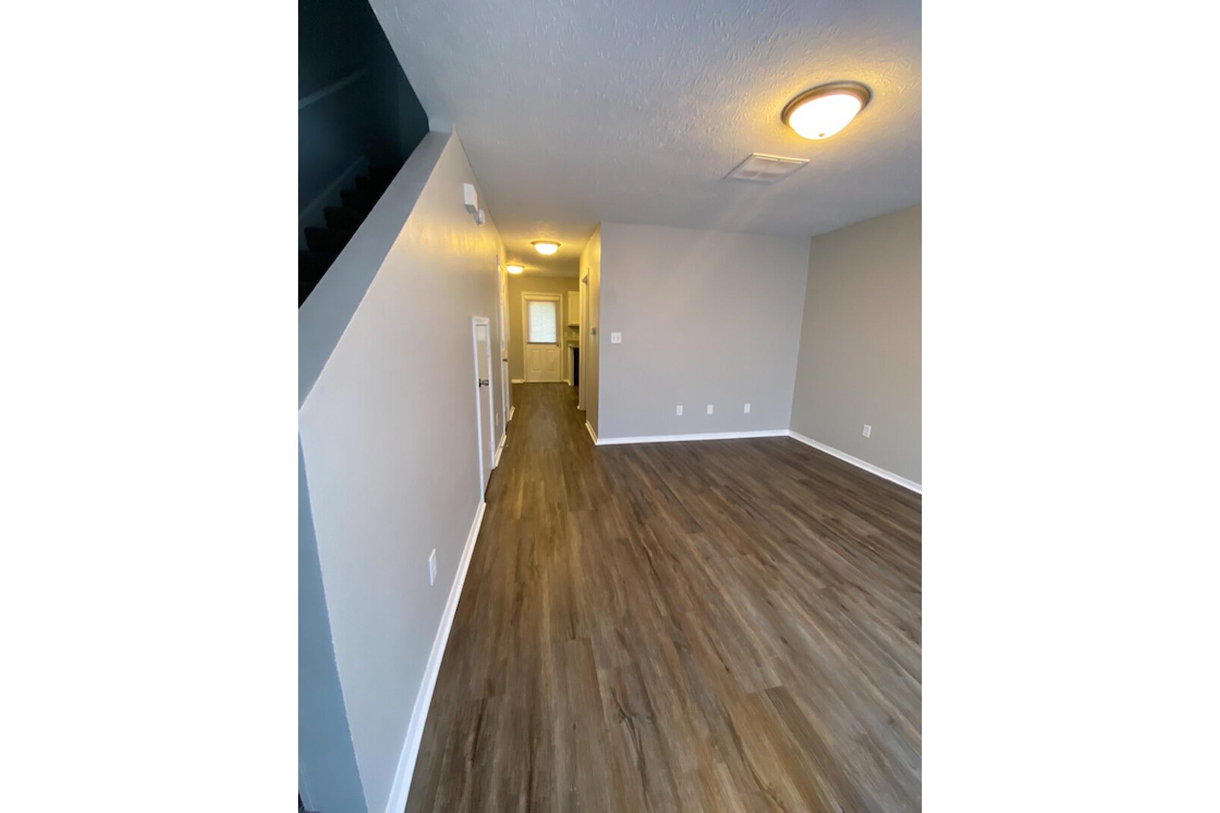 Interior view of a modern living space featuring a long hallway with hardwood flooring, light gray walls, and a bright ceiling light. The hallway leads to a doorway at the end and a staircase on the left. The room is uncluttered and designed in a contemporary style.