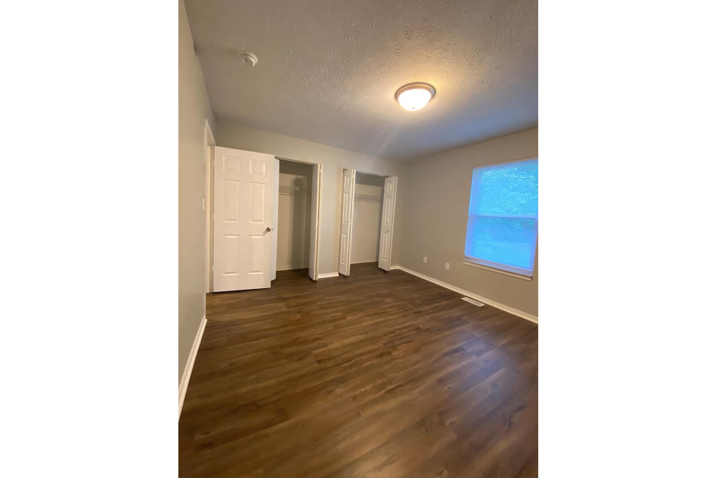 A spacious, empty room with light-colored walls and a hardwood floor. There are two open closet doors on the left side, and a window with blue curtains on the right, letting in natural light. A ceiling light fixture is visible. The overall atmosphere is bright and inviting.