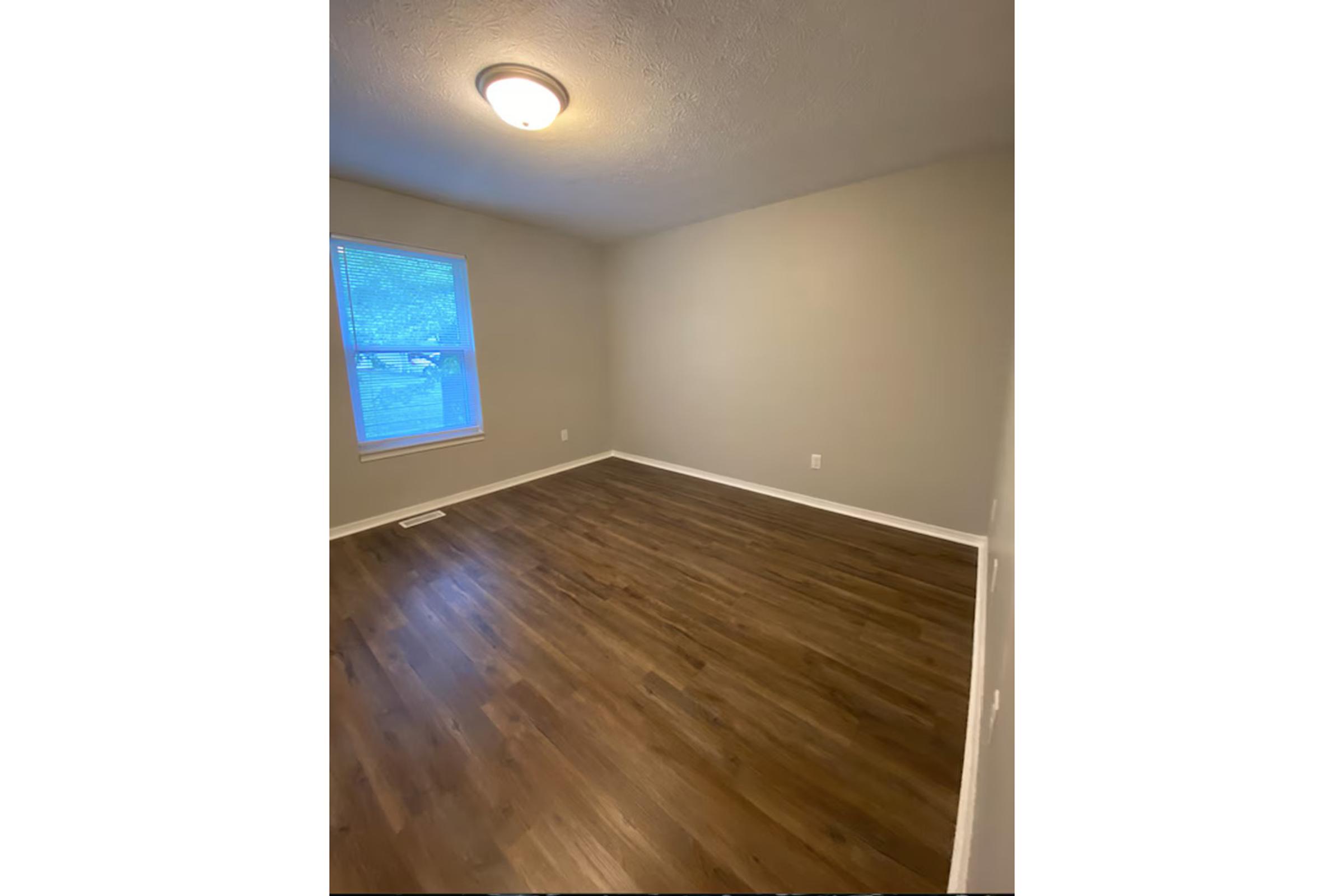 Empty room with light brown wooden flooring, a single window allowing natural light, and a ceiling light fixture. The walls are painted a light beige color. It appears to be a versatile space, suitable for various uses such as a bedroom or office.