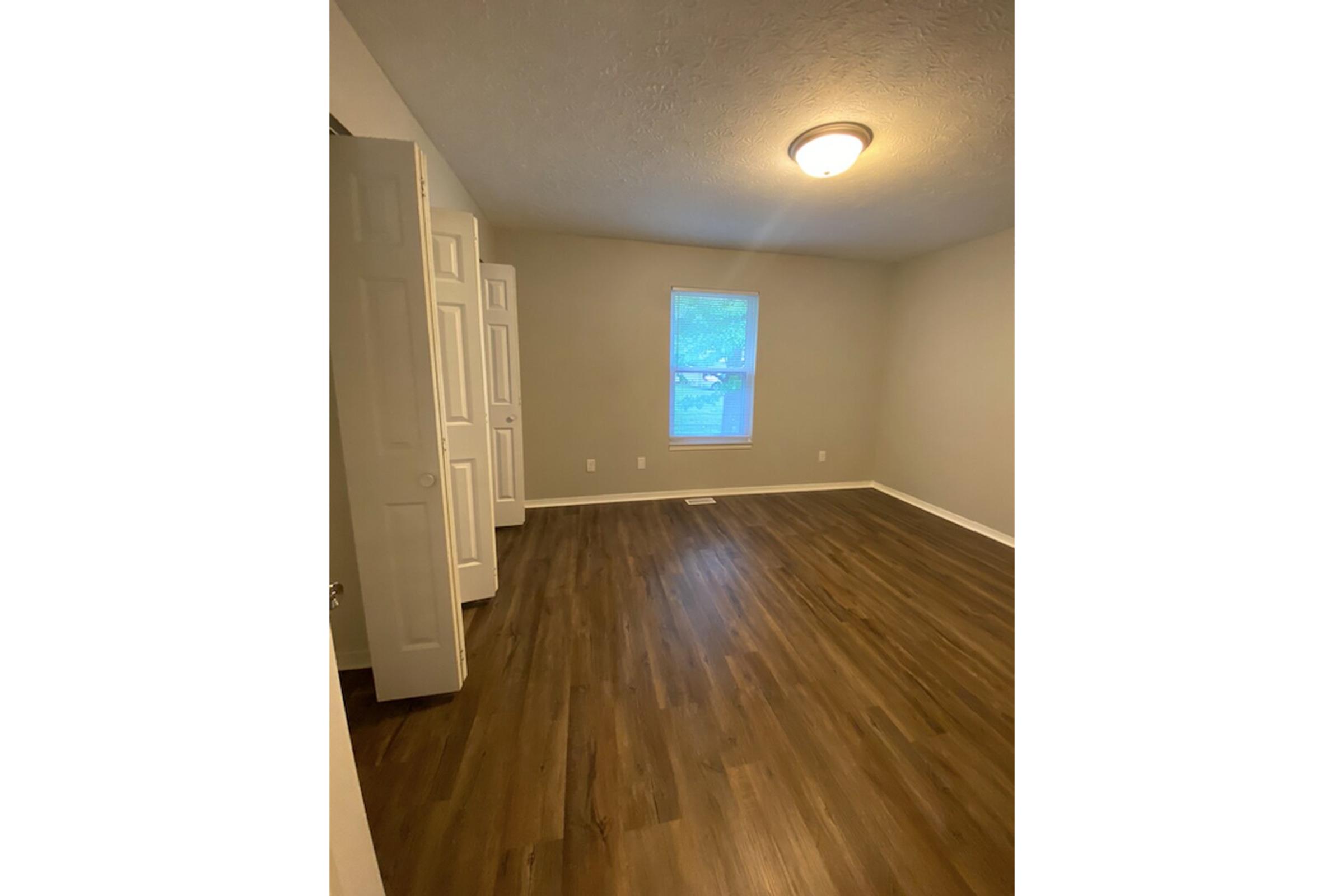 A clean and empty room featuring light-colored walls, a window allowing natural light, and a ceiling light fixture. The floor is made of dark wood laminate, and there are two closed closet doors to the left. The room has a spacious and inviting feel, suitable for various uses.