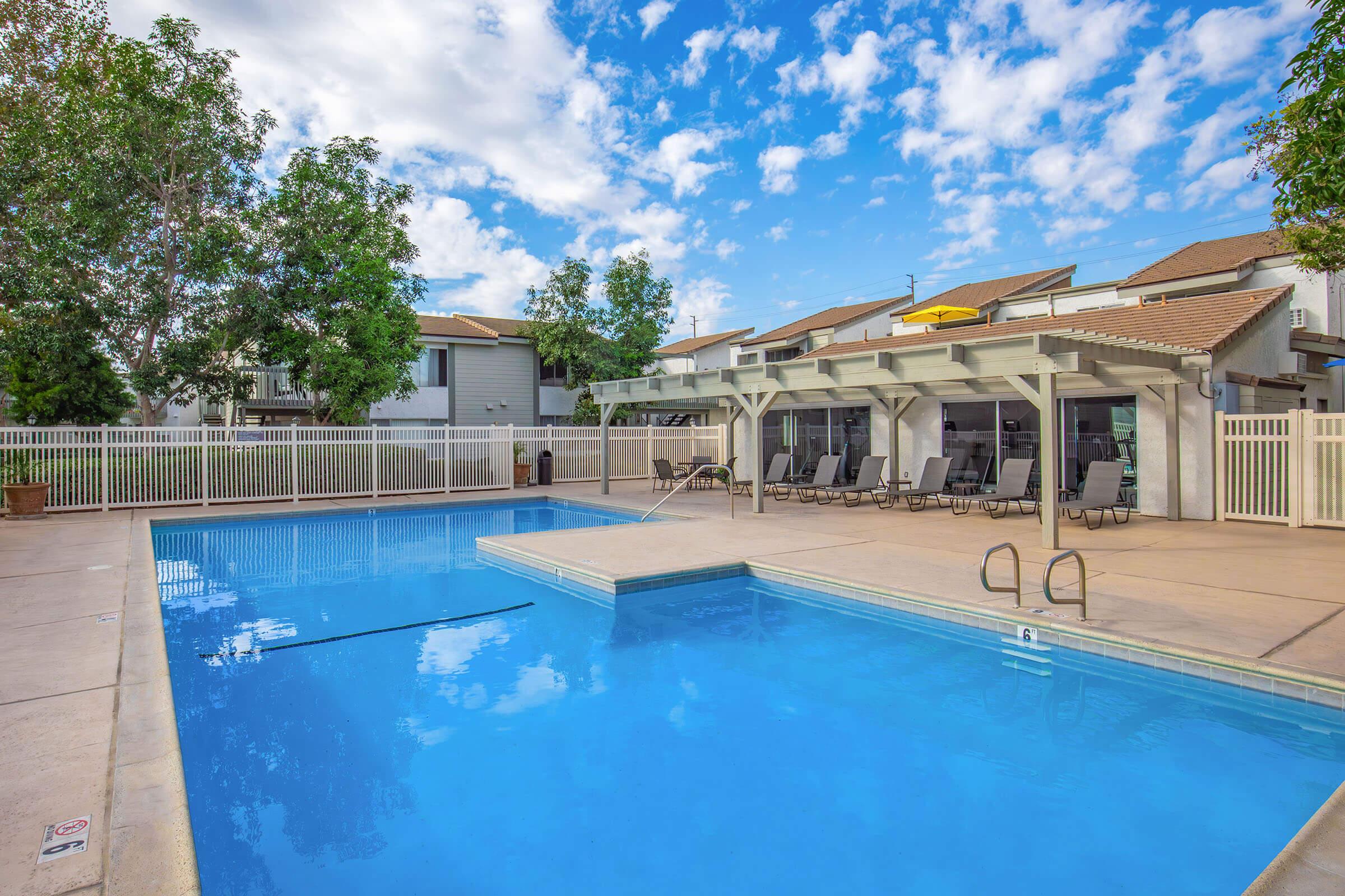 A bright outdoor swimming pool with clear blue water, surrounded by a sunlit patio area. There are lounge chairs under a shaded pergola and a fence lining the area. Lush green trees are visible in the background against a partly cloudy sky, creating a relaxing and inviting atmosphere.
