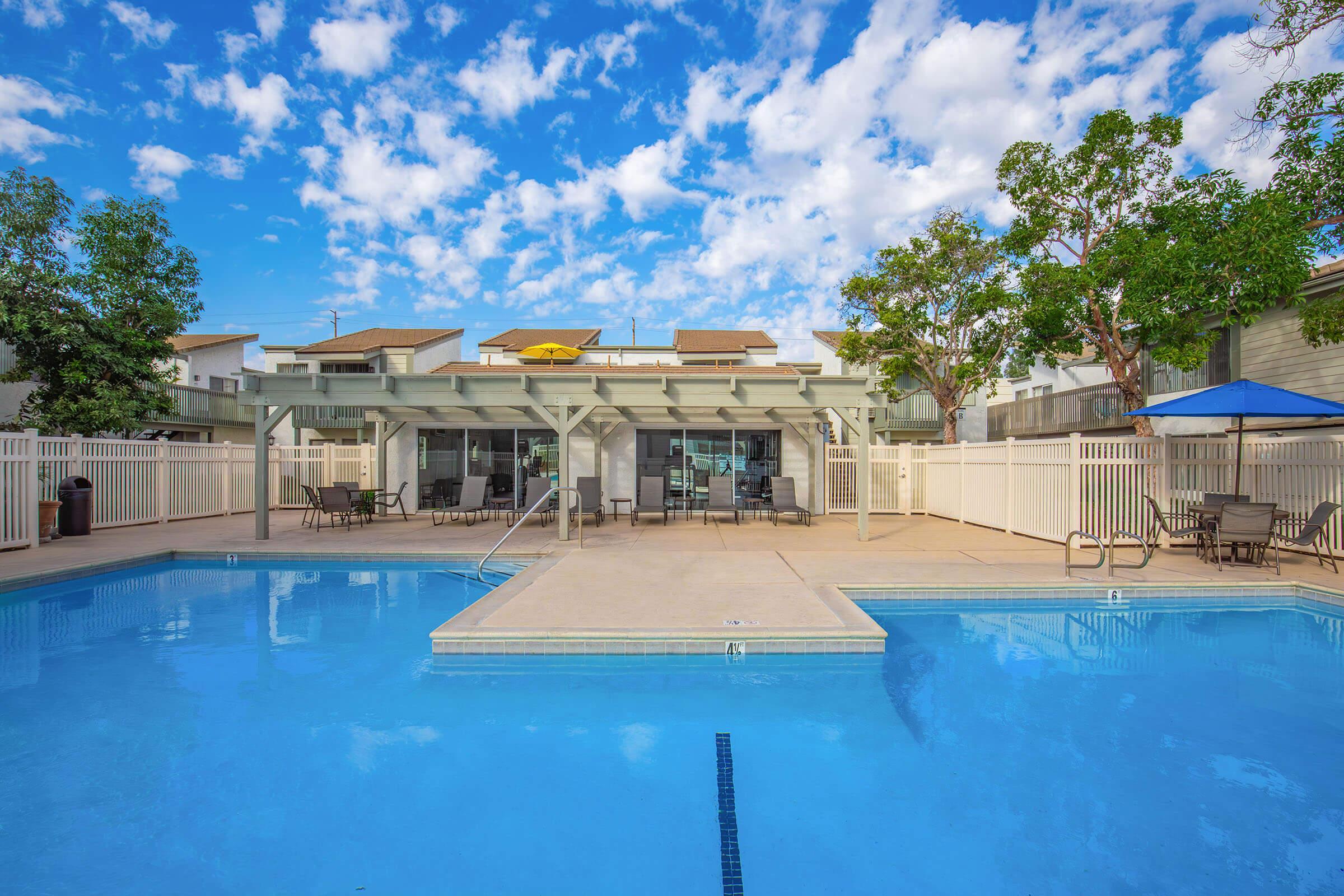 A clear blue swimming pool surrounded by a deck with lounge chairs and tables. In the background, a building with a covered patio area is visible, and colorful umbrellas provide shade. The sky is bright with scattered clouds, creating a sunny atmosphere.