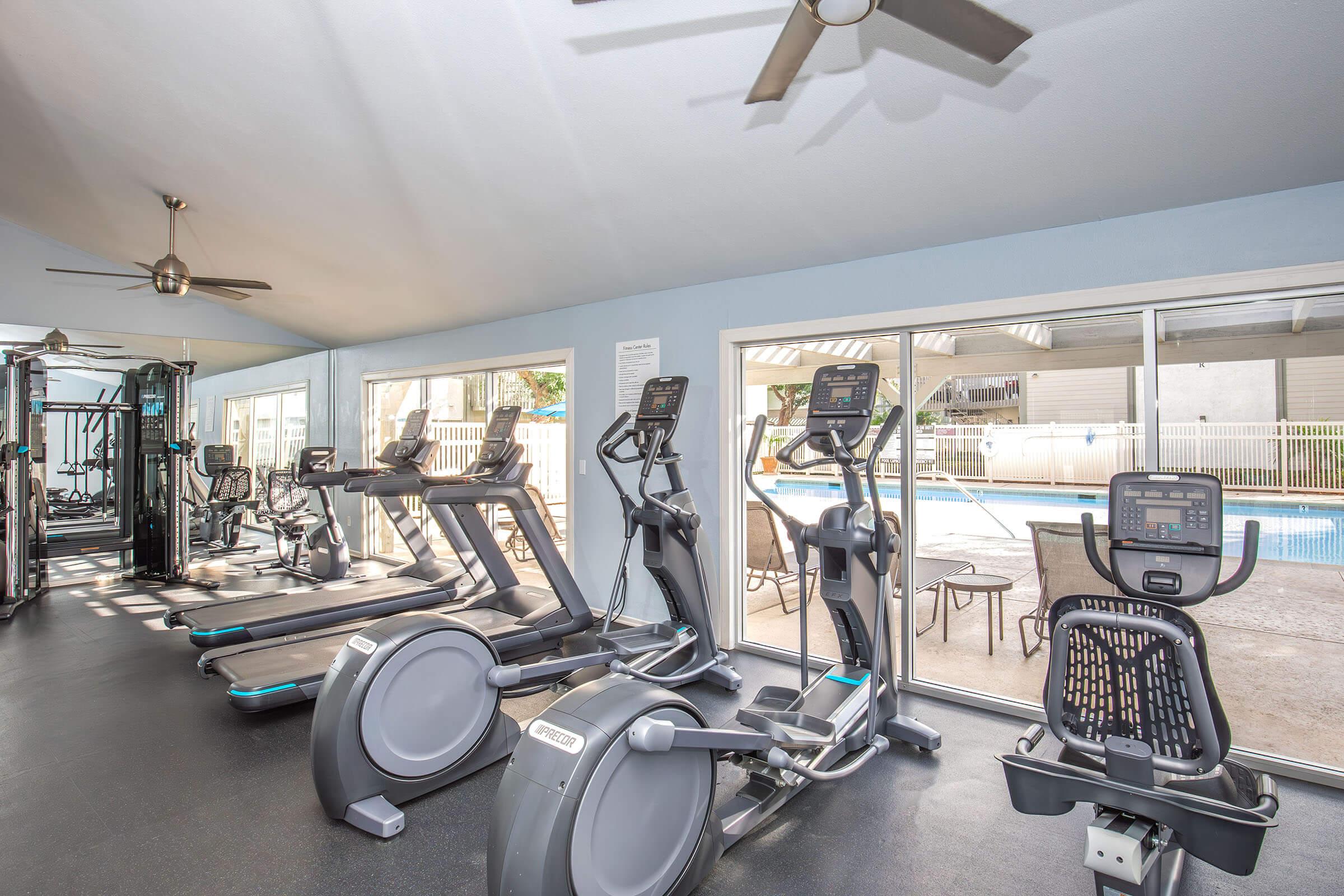 A modern gym interior featuring various exercise equipment, including elliptical machines, stationary bikes, and strength training machines, all positioned near large windows overlooking a swimming pool area. The space is well-lit and has a ceiling fan for ventilation.