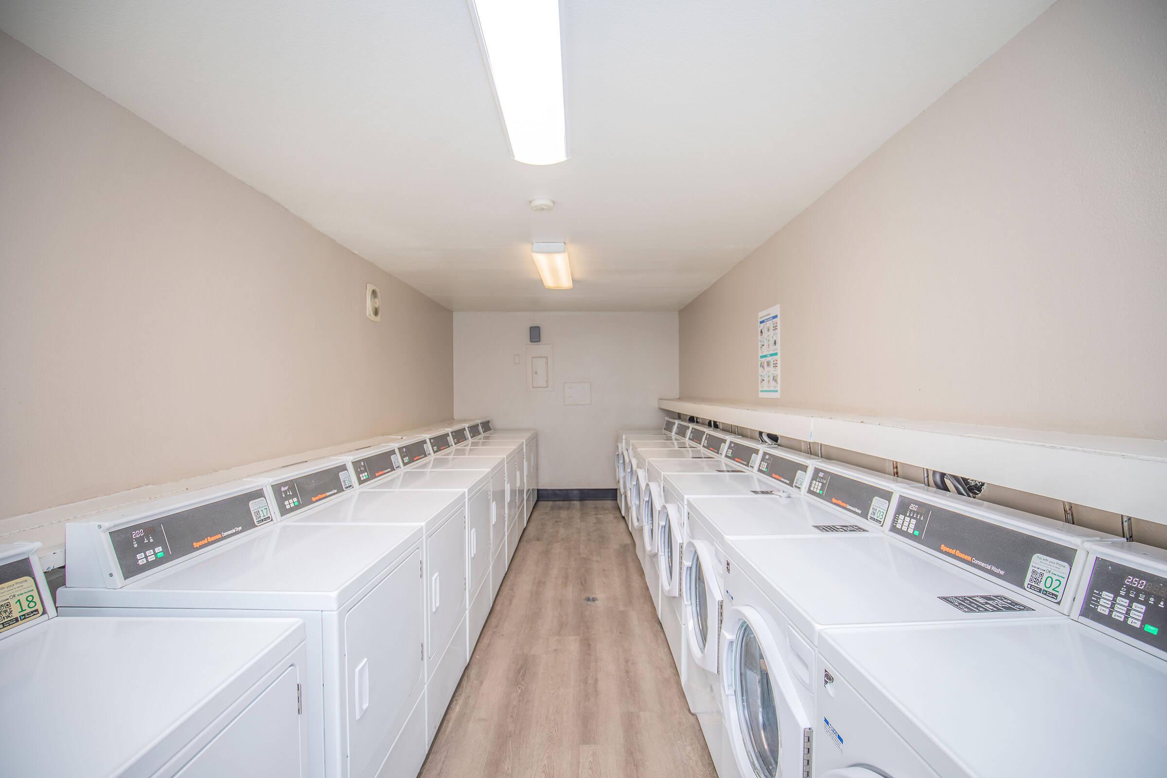 A clean and well-lit laundry room featuring multiple rows of washers and dryers lined against the walls, with light-colored walls and a plain floor. The area is spacious and organized, suitable for laundry tasks. A few informational signs are posted on the walls.
