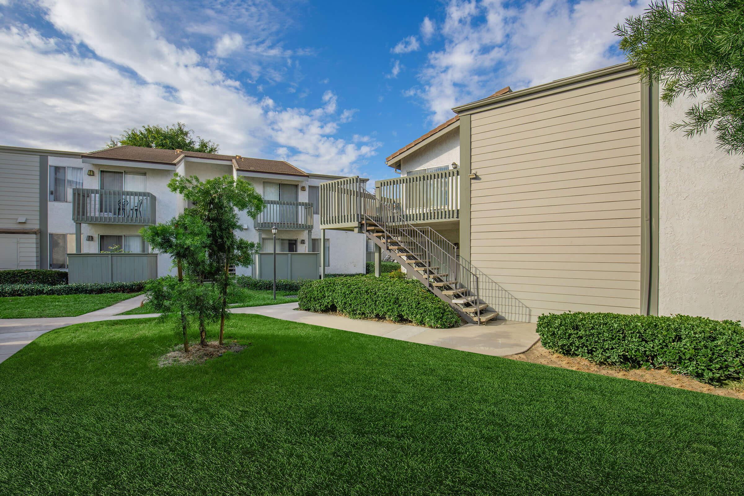 A well-maintained residential area featuring two-story apartment buildings. There are grassy lawns and landscaping with shrubs. A set of stairs leads to the upper level of one building, under a partly cloudy sky. The scene conveys a clean and inviting neighborhood environment.
