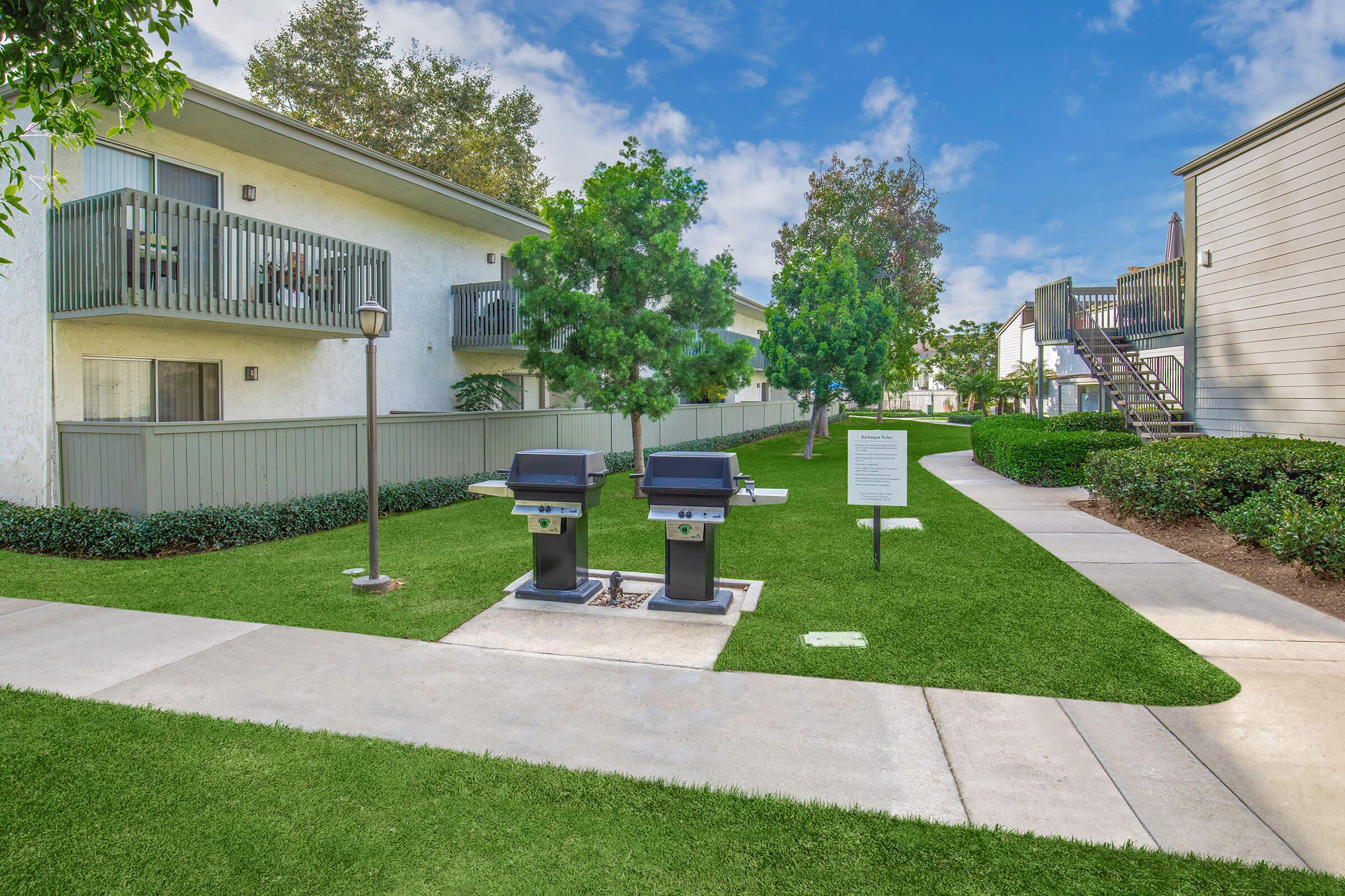 Two modern charcoal grills are positioned on a manicured grassy area surrounded by lush green trees and bushes. A concrete pathway leads to a building with balconies, and there are additional steps leading up to another building in the background. The sky is clear with a few clouds.
