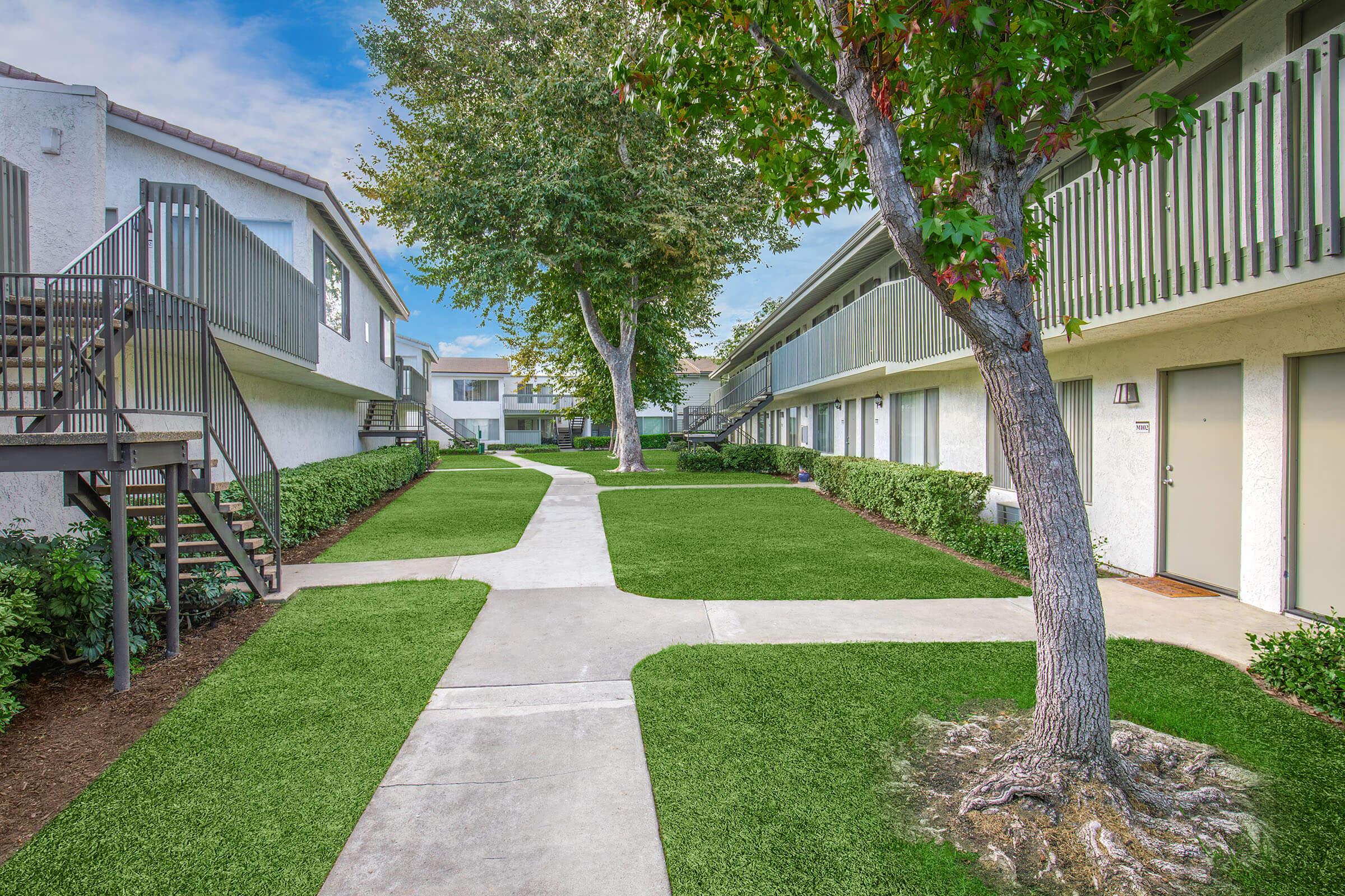 A well-maintained outdoor courtyard with manicured green lawns, concrete pathways, and apartment buildings on either side. There are trees providing shade, and a staircase leading to a second-floor apartment. The scene is bright and inviting, suggesting a pleasant living environment.