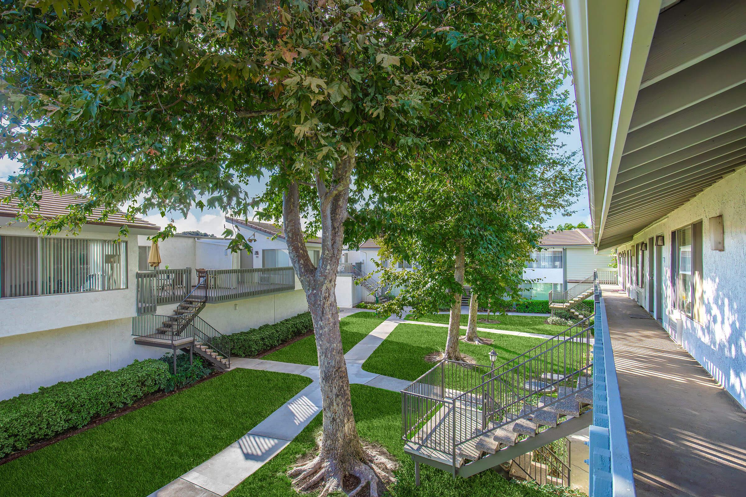A landscaped area of a residential complex featuring green grass and trees. There are pathways and stairs leading to different units, with a covered walkway on one side. The setting is bright and well-maintained, creating a serene environment.