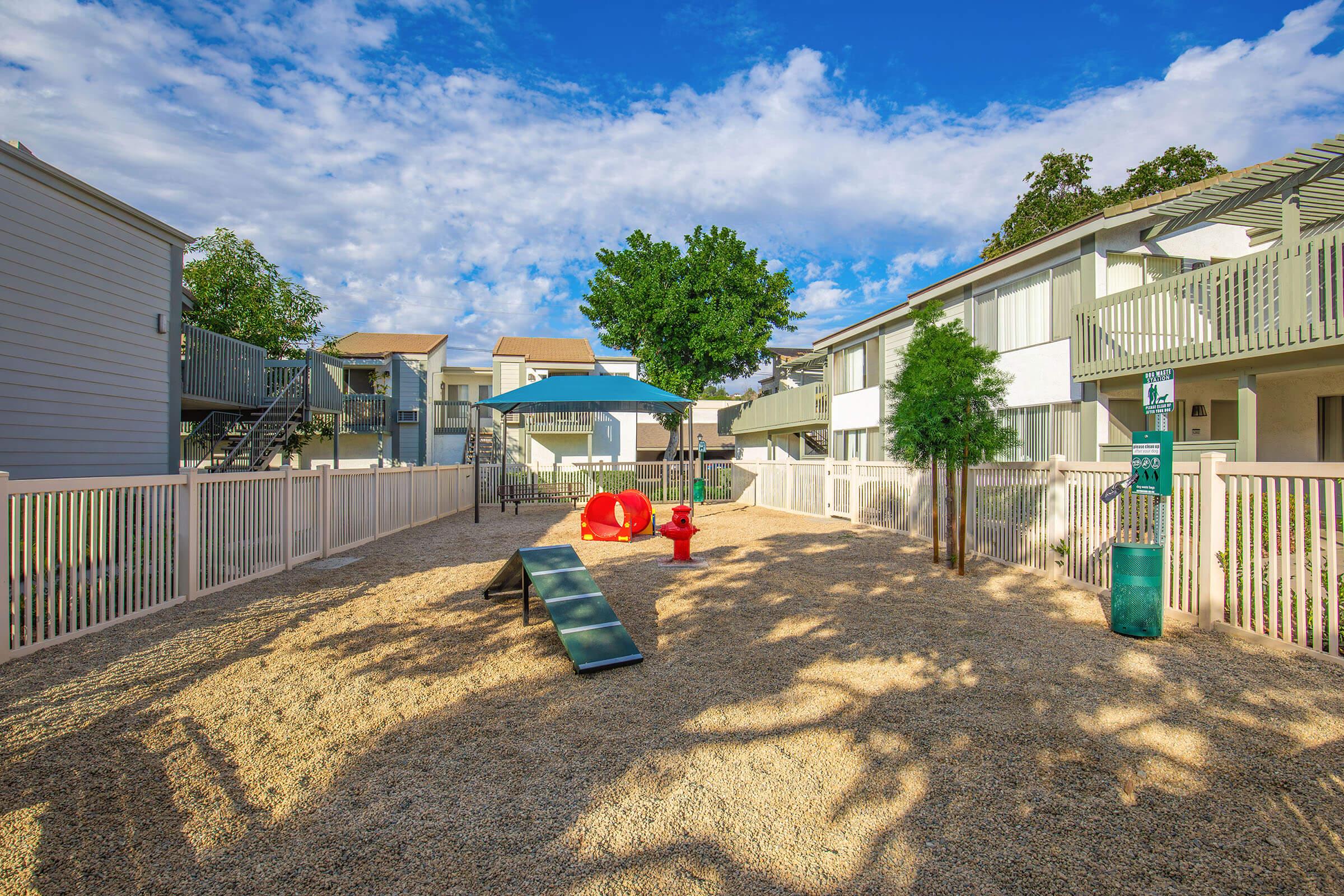 A playground area featuring a red tunnel slide, a climbing structure, and a sand-filled space, surrounded by white fencing. Nearby, there are trees and two residential buildings under a blue sky with scattered clouds. The setting is bright and inviting, suitable for children.
