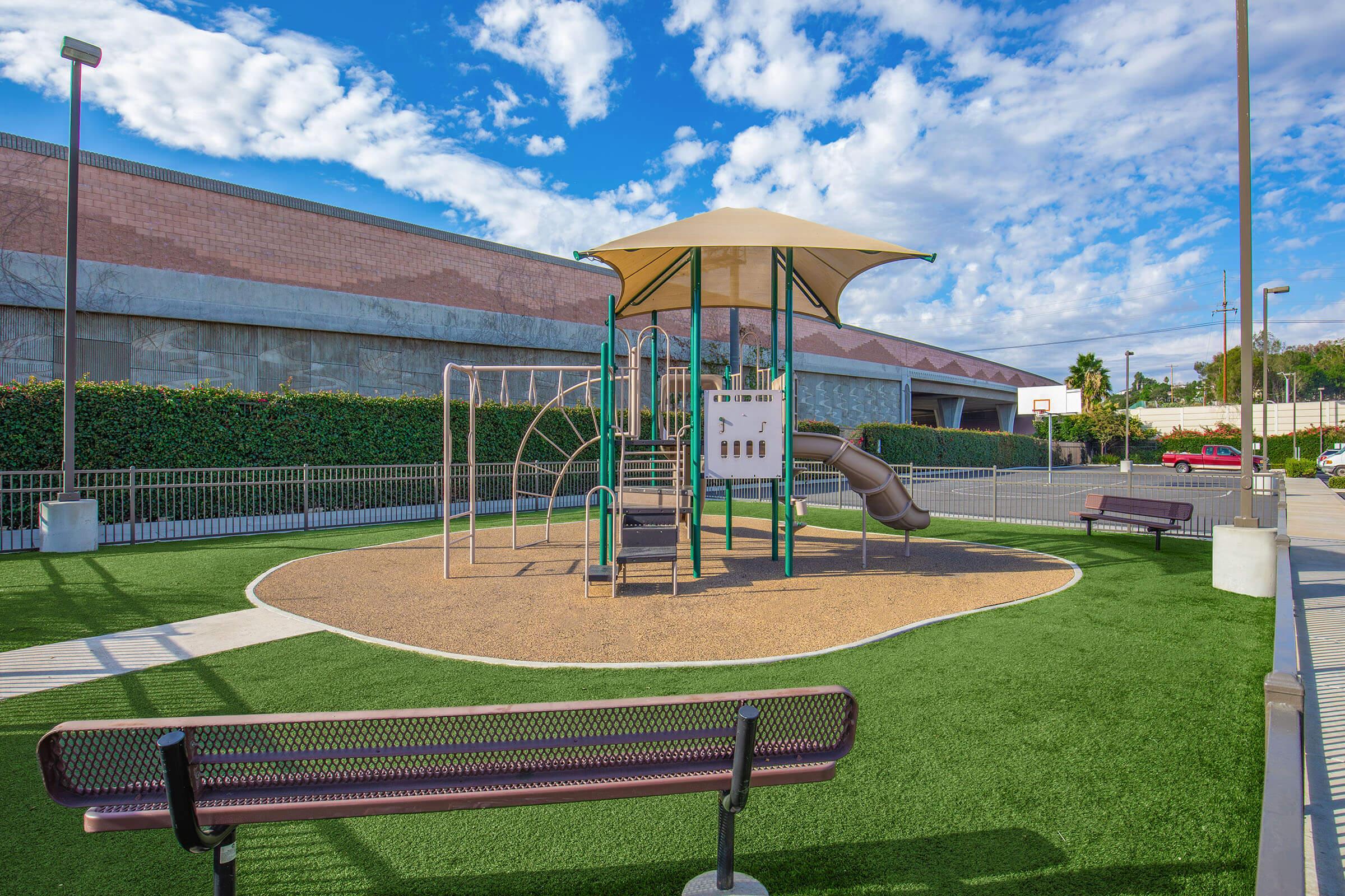 A playground with a climbing structure and slide, surrounded by green grass and benches. The area is sunny with a partly cloudy sky in the background, and there are walls and trees nearby. The playground is empty, inviting play and activity.